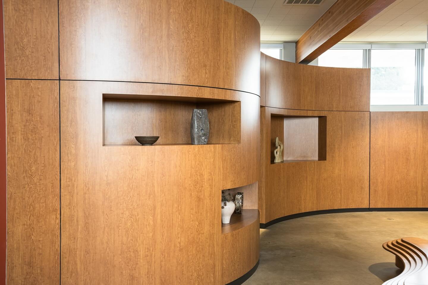 Interior of a room with curved wooden wall shelving displaying decorative objects and a curved bench on the floor.