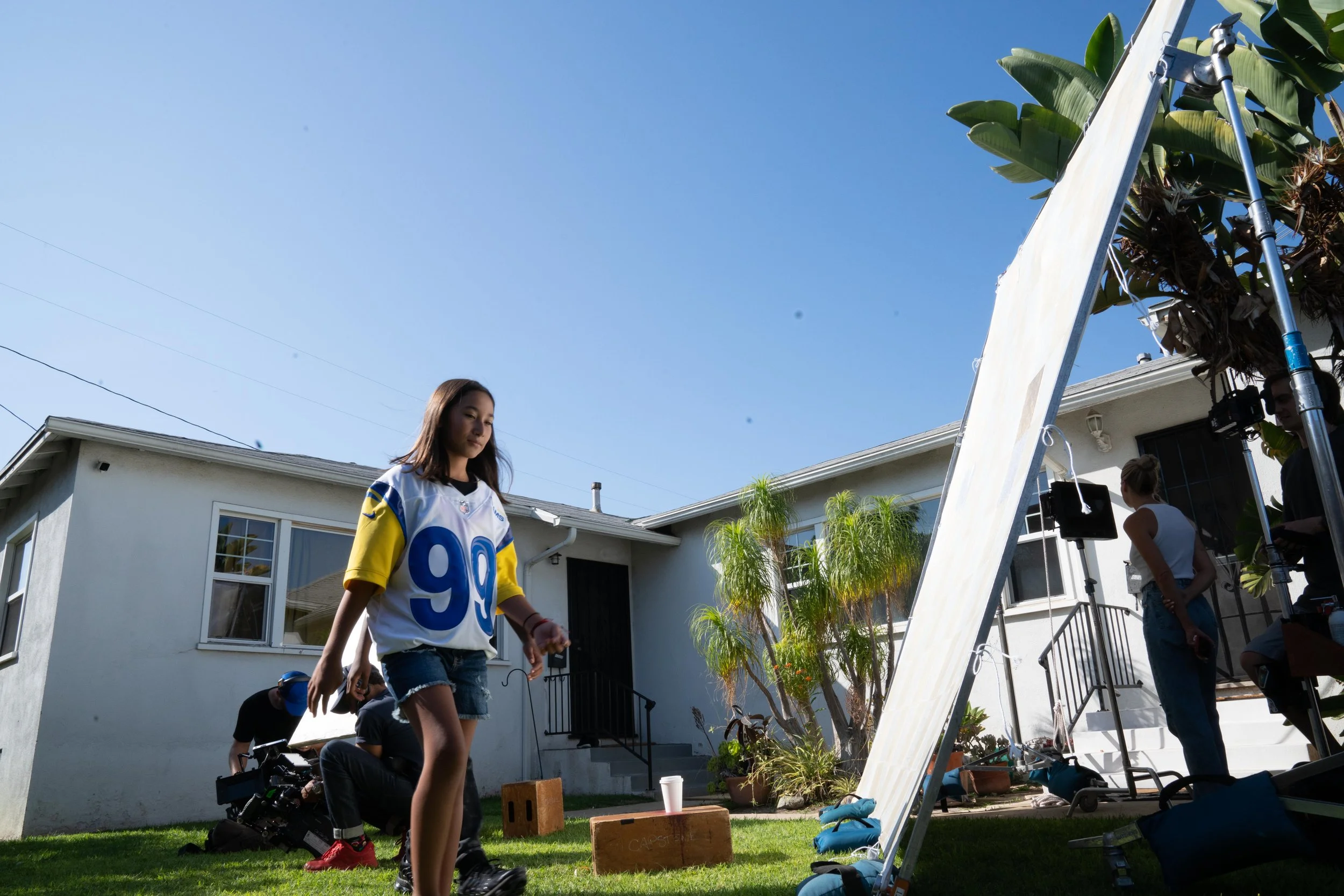 Girl walks on a lawn during behind-the-scenes filming with crew members and equipment, house and blue sky in background.