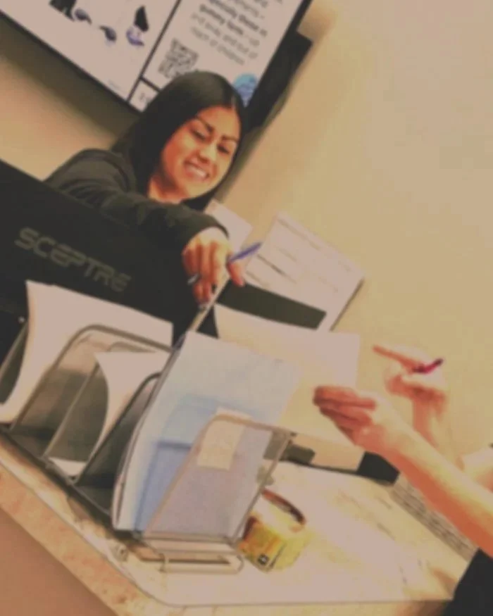 A woman smiling and reaching out to shake hands with another person in an office or classroom setting.