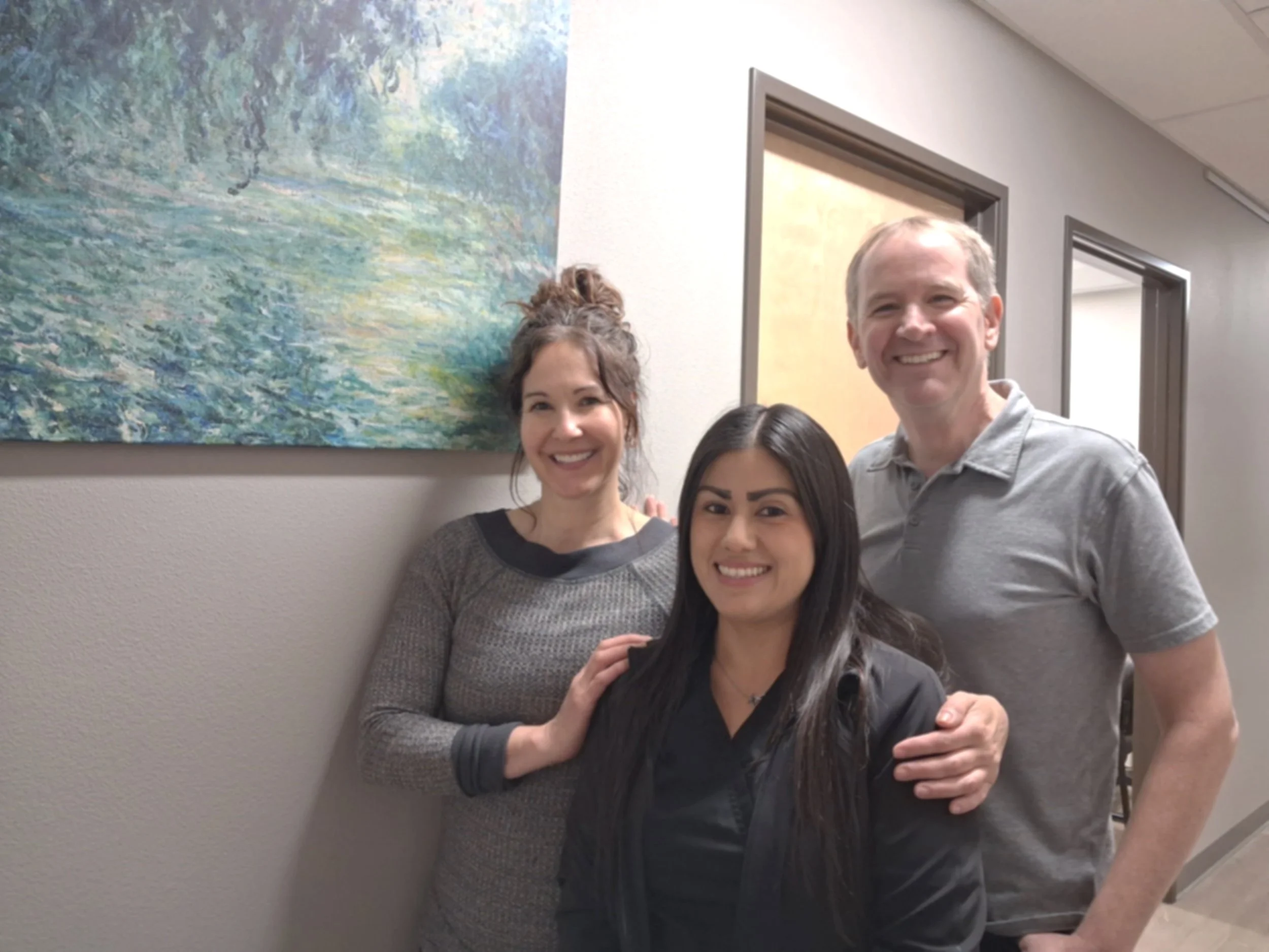 Three people smiling indoors, standing near a beige wall with a colorful abstract painting behind them.