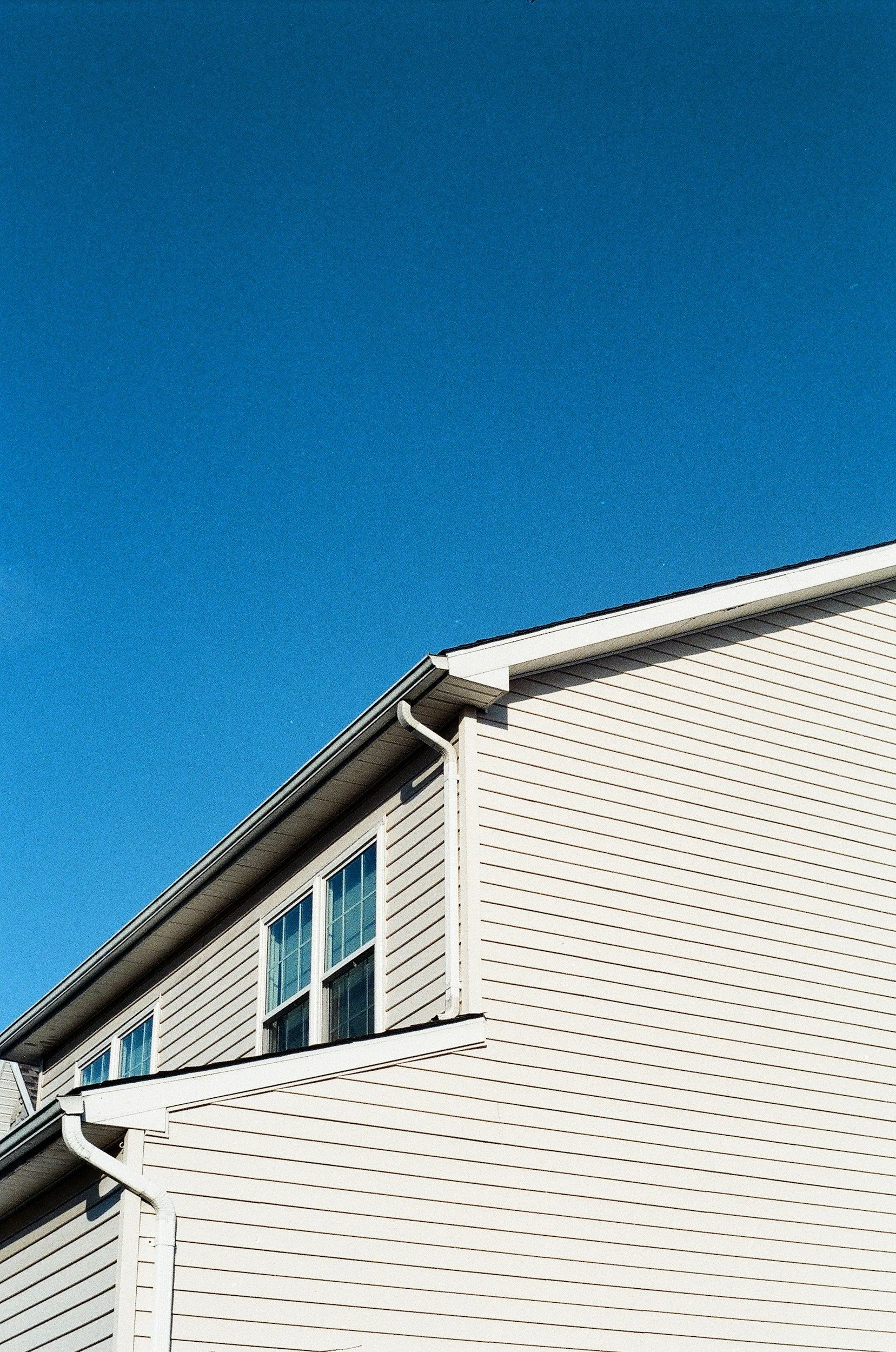 Close-up of the upper corner of a beige house with white trim, two windows, and a clear blue sky in the background.