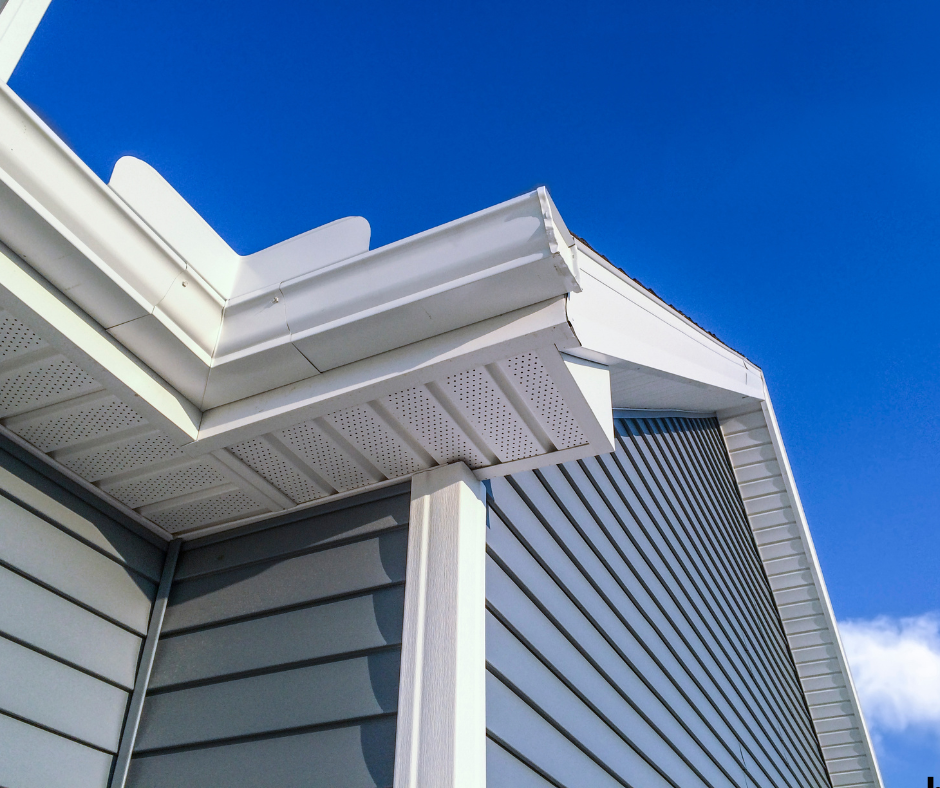 Close-up of the corner of a house roof with white gutter and siding against a bright blue sky.