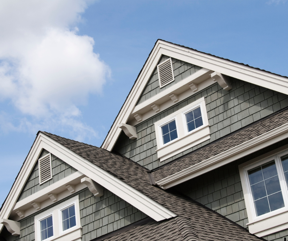 Close-up of the upper part of a gray house with shingle siding, white window frames, and gabled roof against a blue sky with clouds.