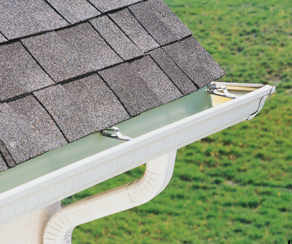Close-up of a house's roof showing asphalt shingles and a gutter system with a downspout, overlooking a grassy area.