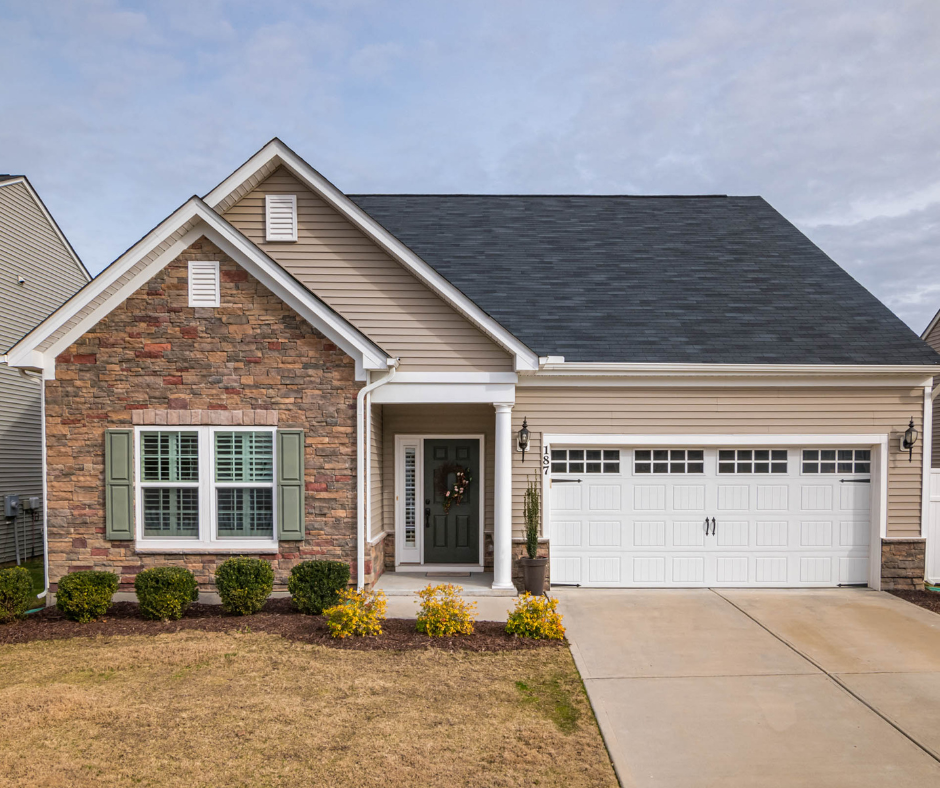 Front view of a suburban house with a brick and siding exterior, green shutters, a gable roof, a two-car garage, a front yard with bushes and small yellow flowers, and a front porch with a decorated door.