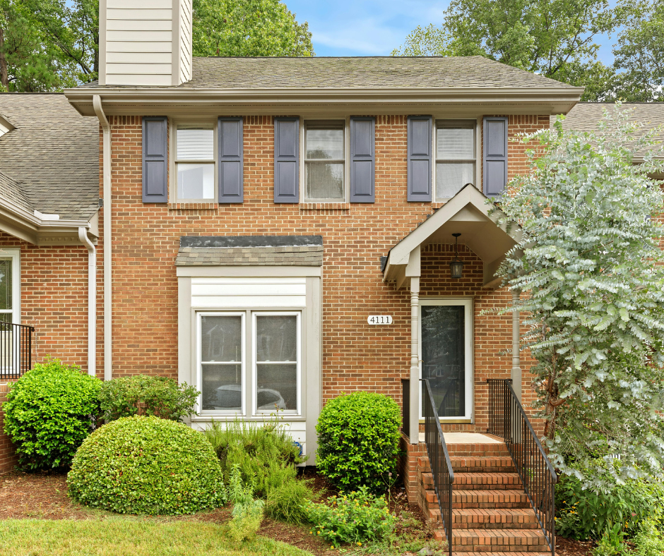 Front view of a two-story brick house with blue shutters and a small front porch with stairs and black railing, surrounded by green shrubs and trees