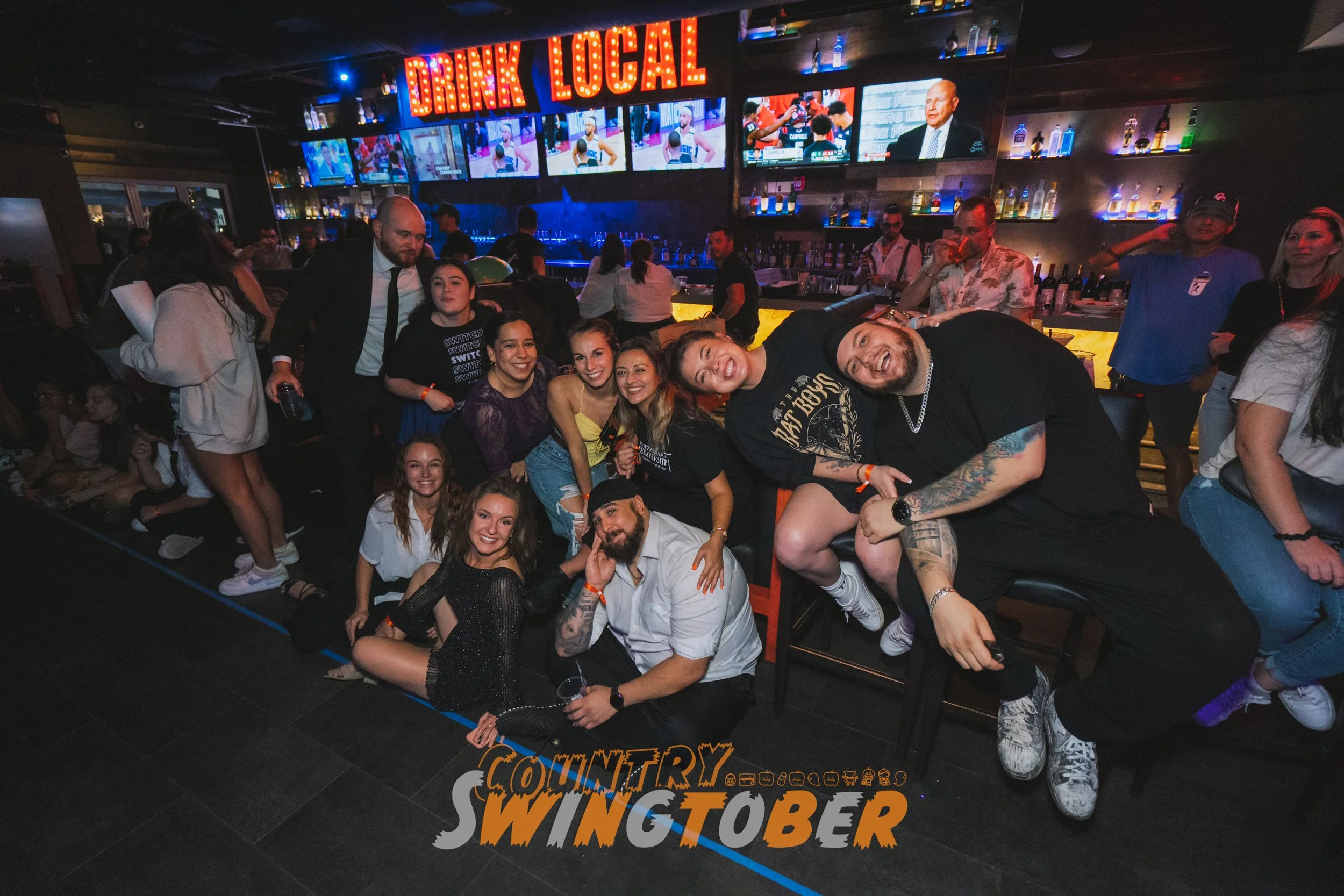 A group of people enjoying a night out at a bar with multiple TV screens and a neon sign that reads 'Drink Local.' The group is posing together happily, some sitting on the floor and others standing or sitting on chairs, with a lively bar scene in the background.