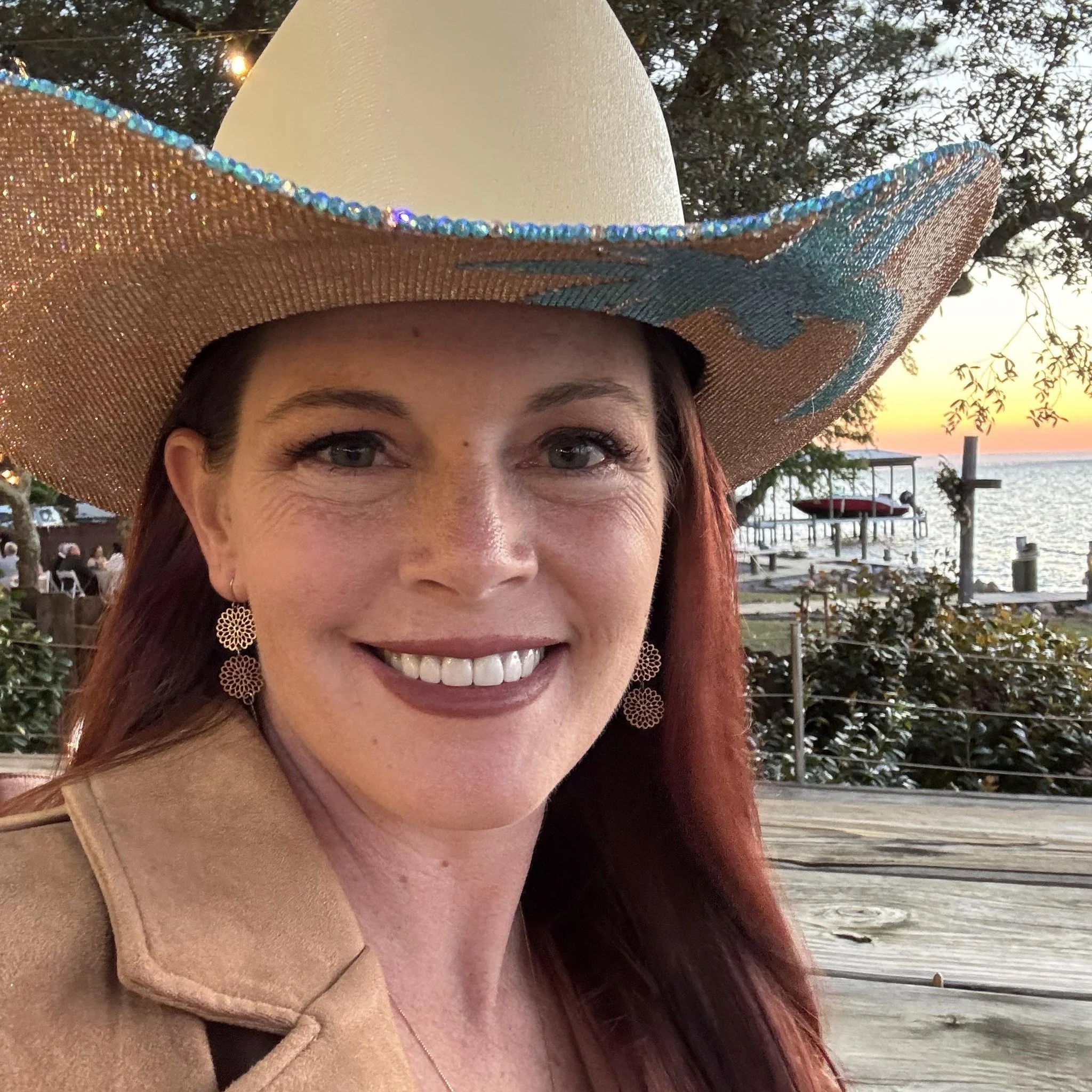 A woman with red hair smiling at the camera, wearing a large cowboy hat with decorative beads, earrings, and a tan coat. In the background, there is a sunset over a body of water with a dock, trees, and boats.