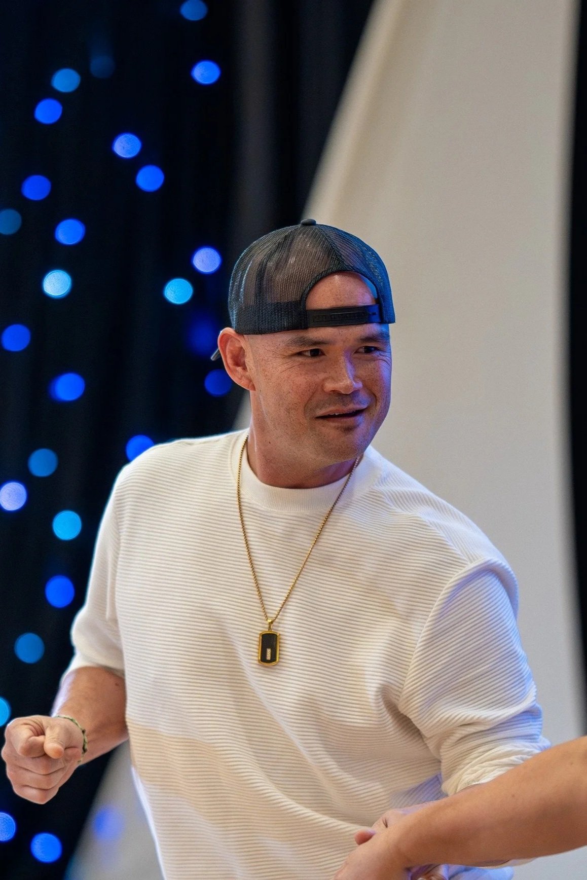 A man wearing a white shirt, a black baseball cap backward, and a gold necklace with a pendant, smiling and holding hands with someone off-camera, with blue string lights in the background.