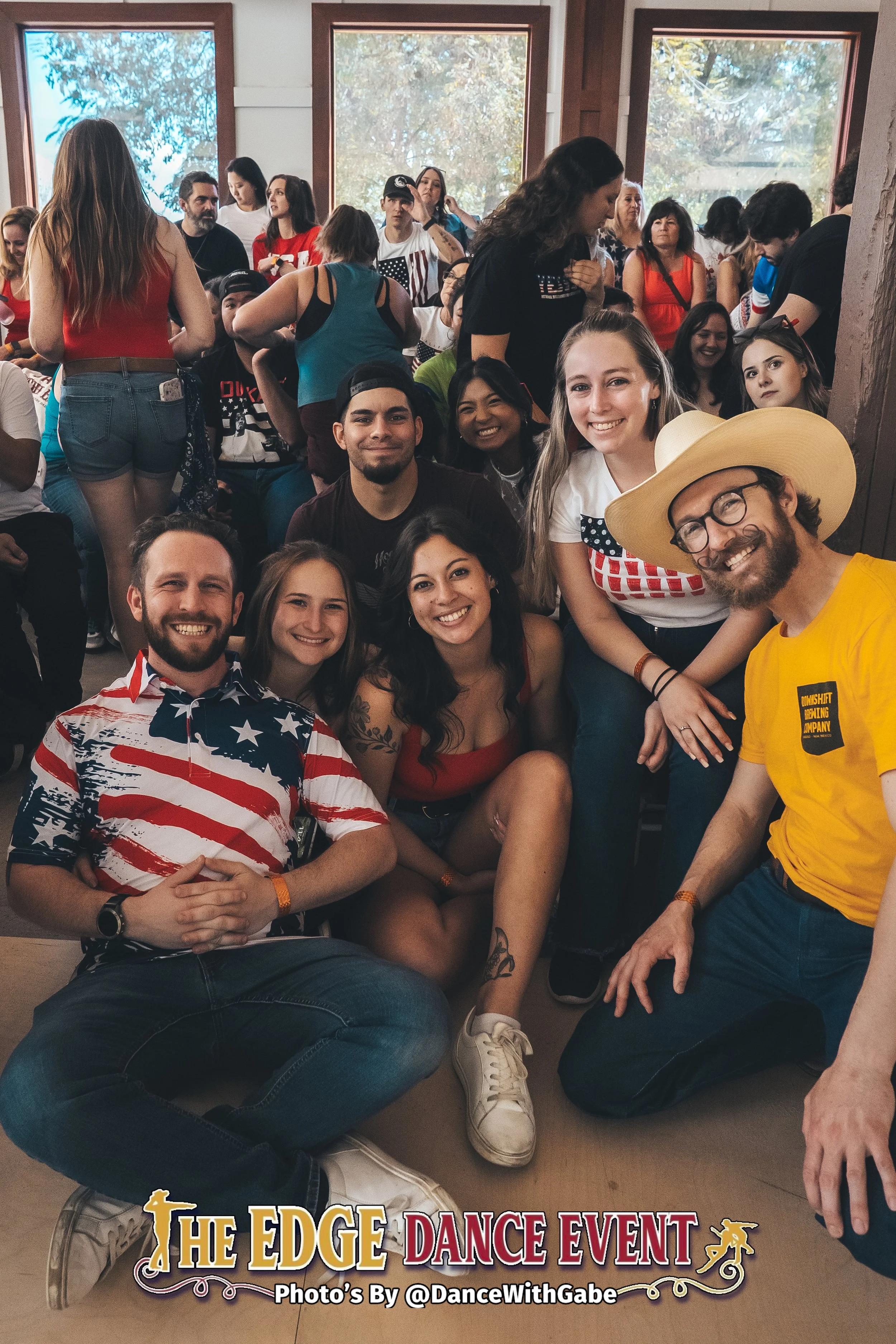A group of smiling young adults gathered together at a dance event, with some sitting and others standing in a crowded room, celebrating at The Edge Dance Event.