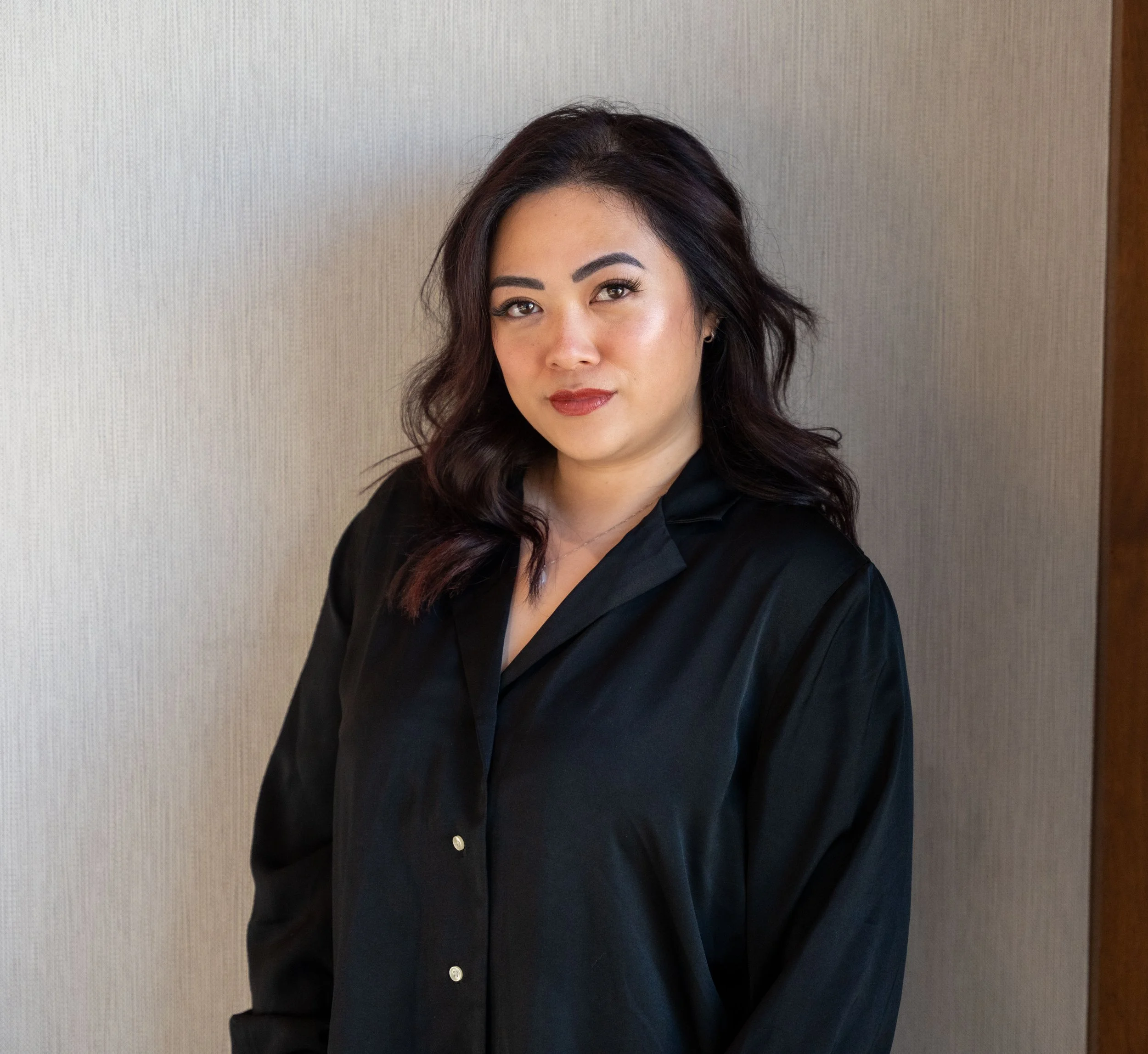 A woman with dark wavy hair, wearing makeup, and a black blouse standing against a neutral-colored wall.