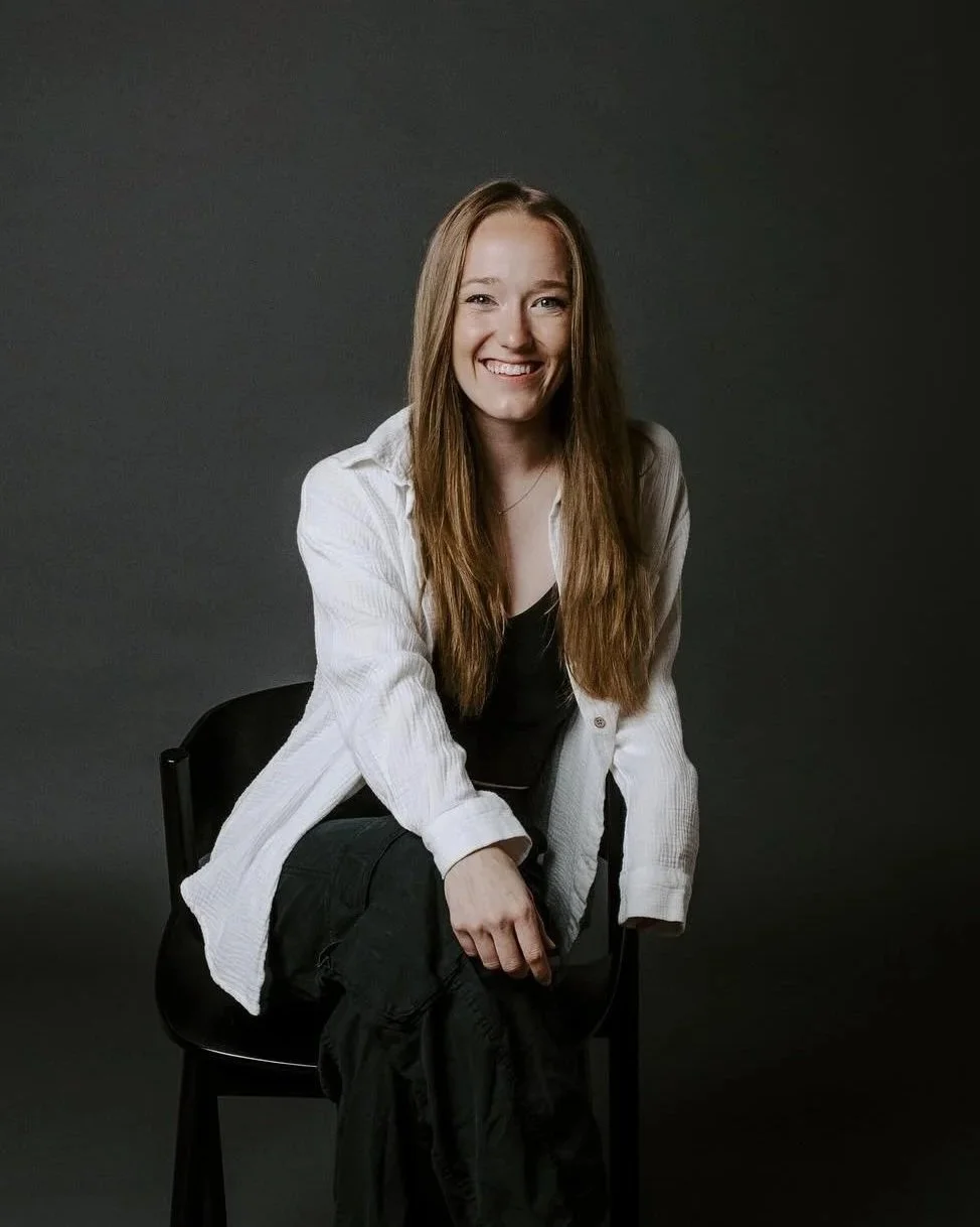 A young woman with long red hair, smiling, wearing a white button-up shirt over a black top, sitting on a black chair against a plain dark background.