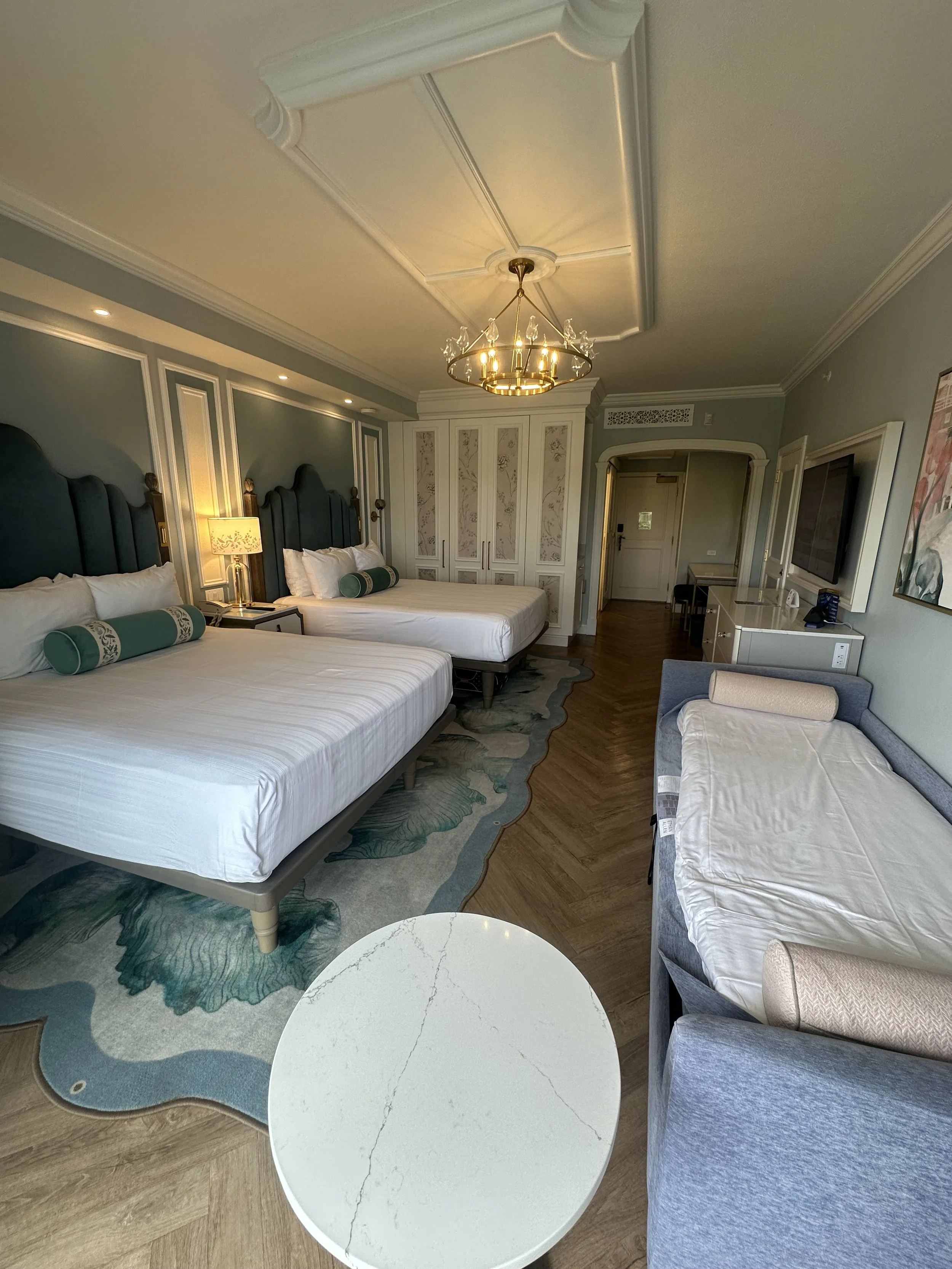 Interior of a hotel room with two large beds, a chandelier, a flat-screen TV, a white round table, and a daybed.