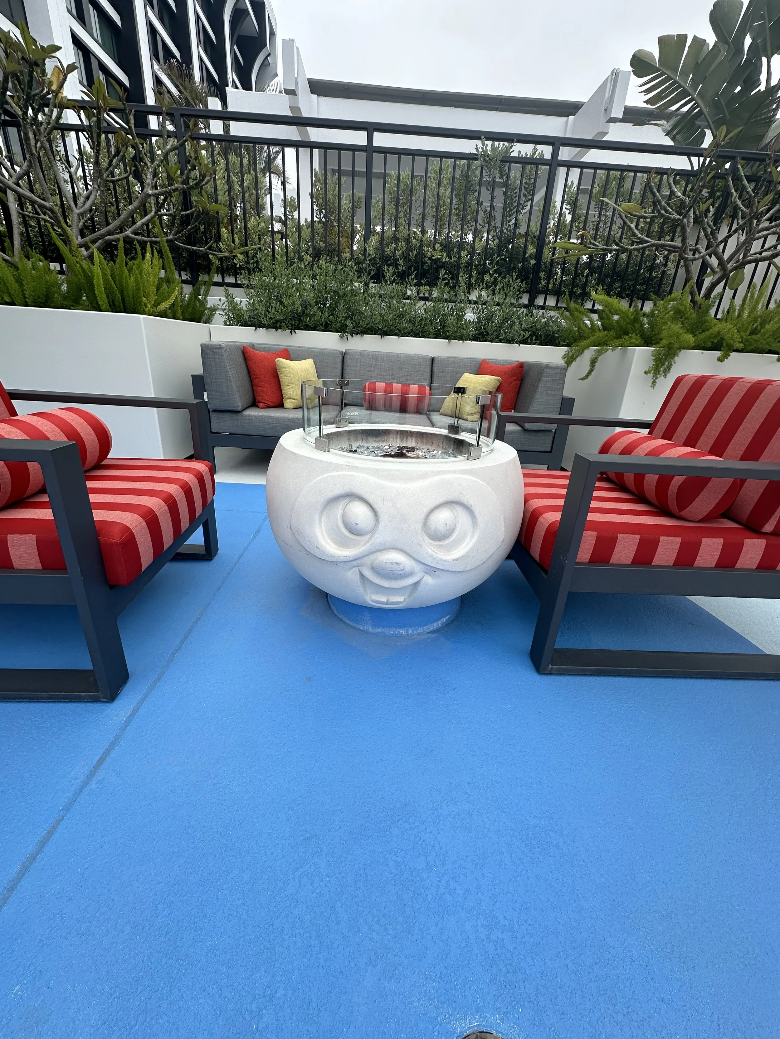 Outdoor patio with a white ceramic fire pit in the shape of a face, surrounded by three chairs with red and striped cushions, and a gray couch with colorful pillows, set on a blue floor with greenery and a black railing in the background.