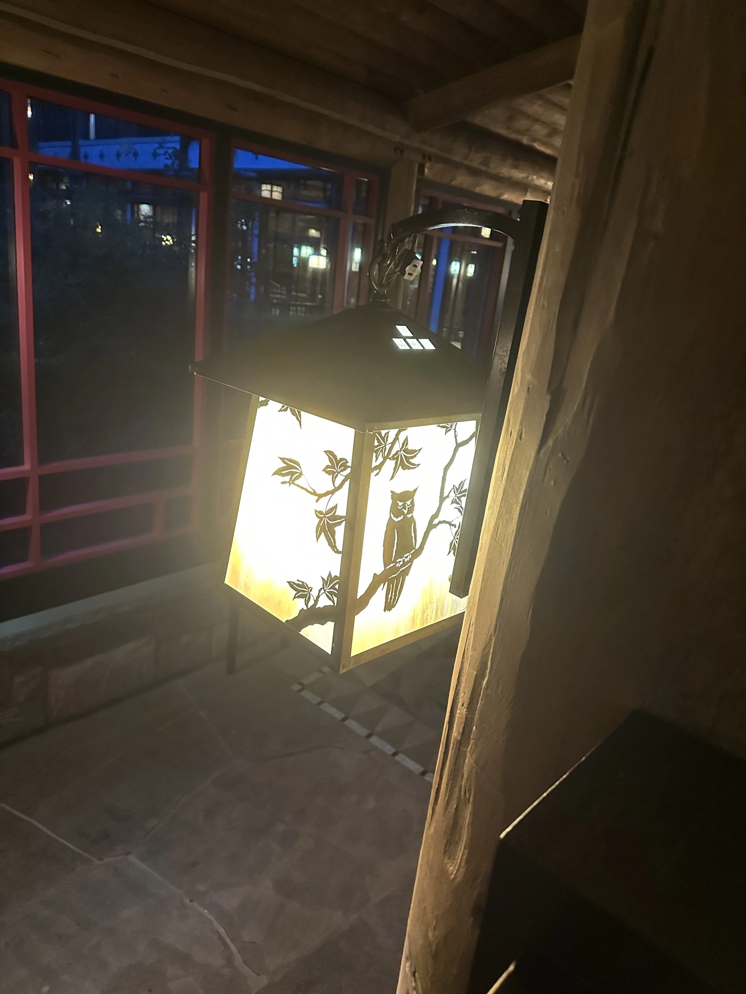 A decorative illuminated lantern with owl and branch design inside a wooden structure near a window at night.