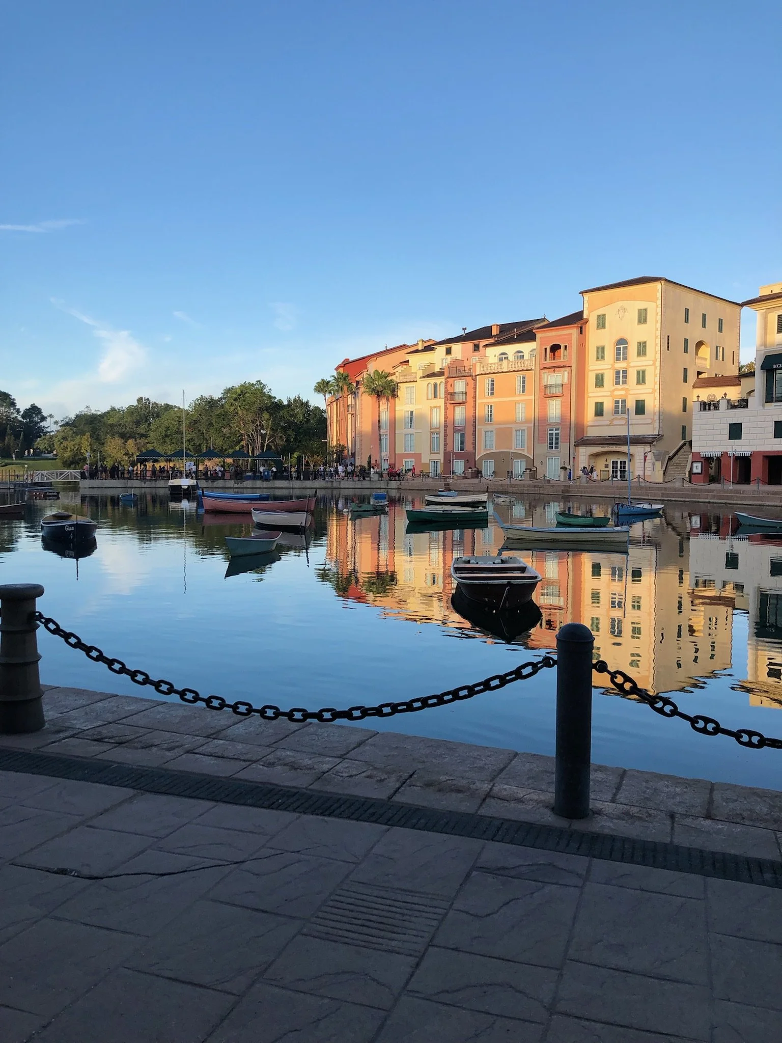 Colorful buildings along a waterfront with boats floating in the water, reflections visible on the surface, and clear blue sky.
