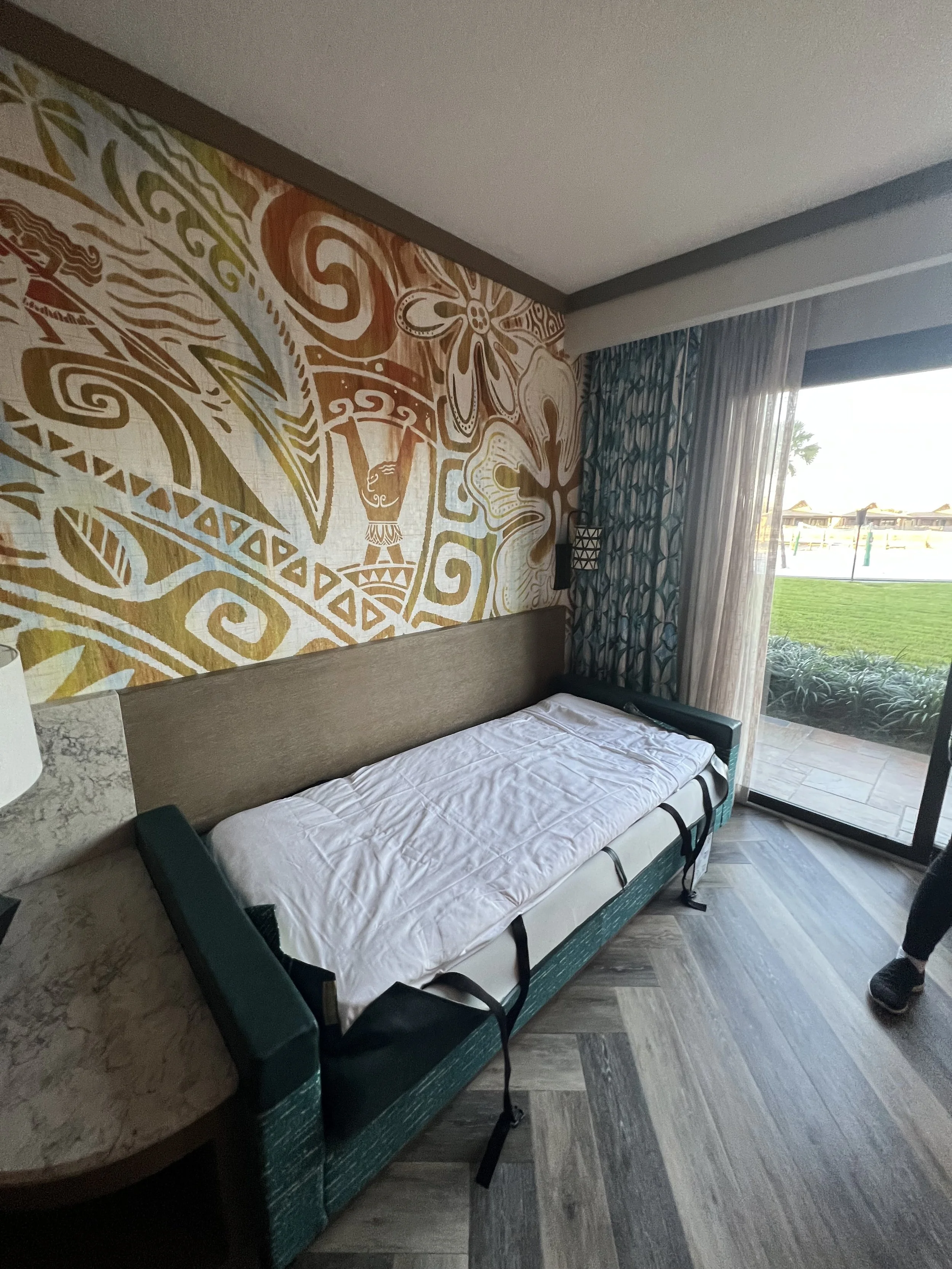 A hotel room with a sofa bed covered with a folded white sheet, a wall with a colorful tribal-patterned mural, a sliding glass door with sheer curtains, and a view of a green lawn outside.