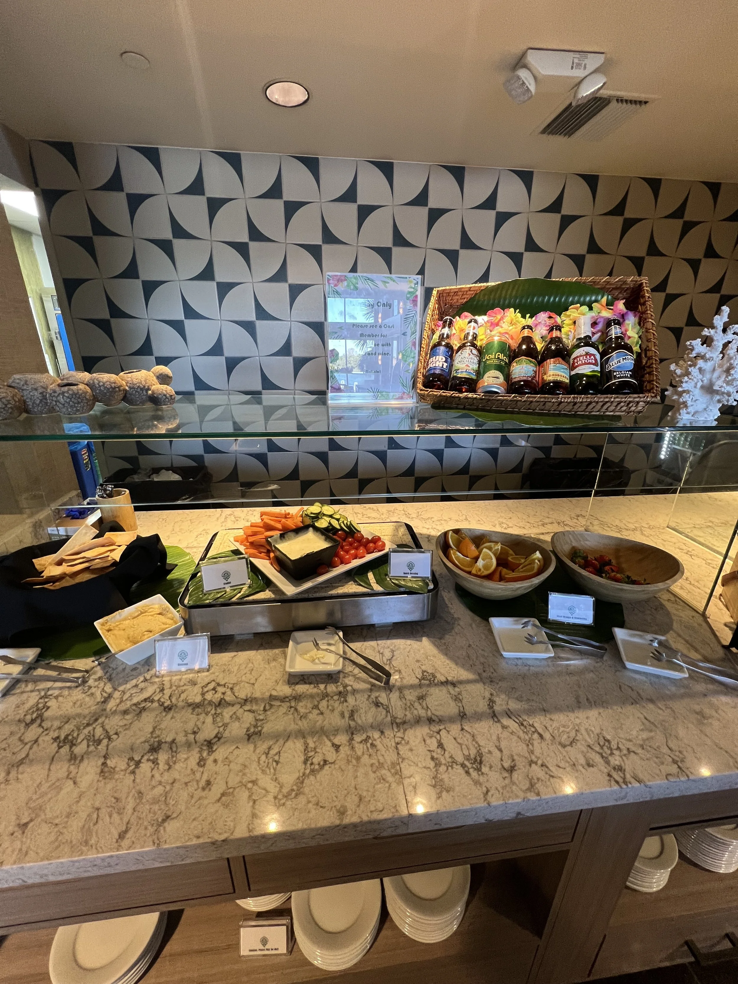 Buffet spread with vegetables, sliced fruits, condiments, and beverages on a marble countertop in a restaurant.