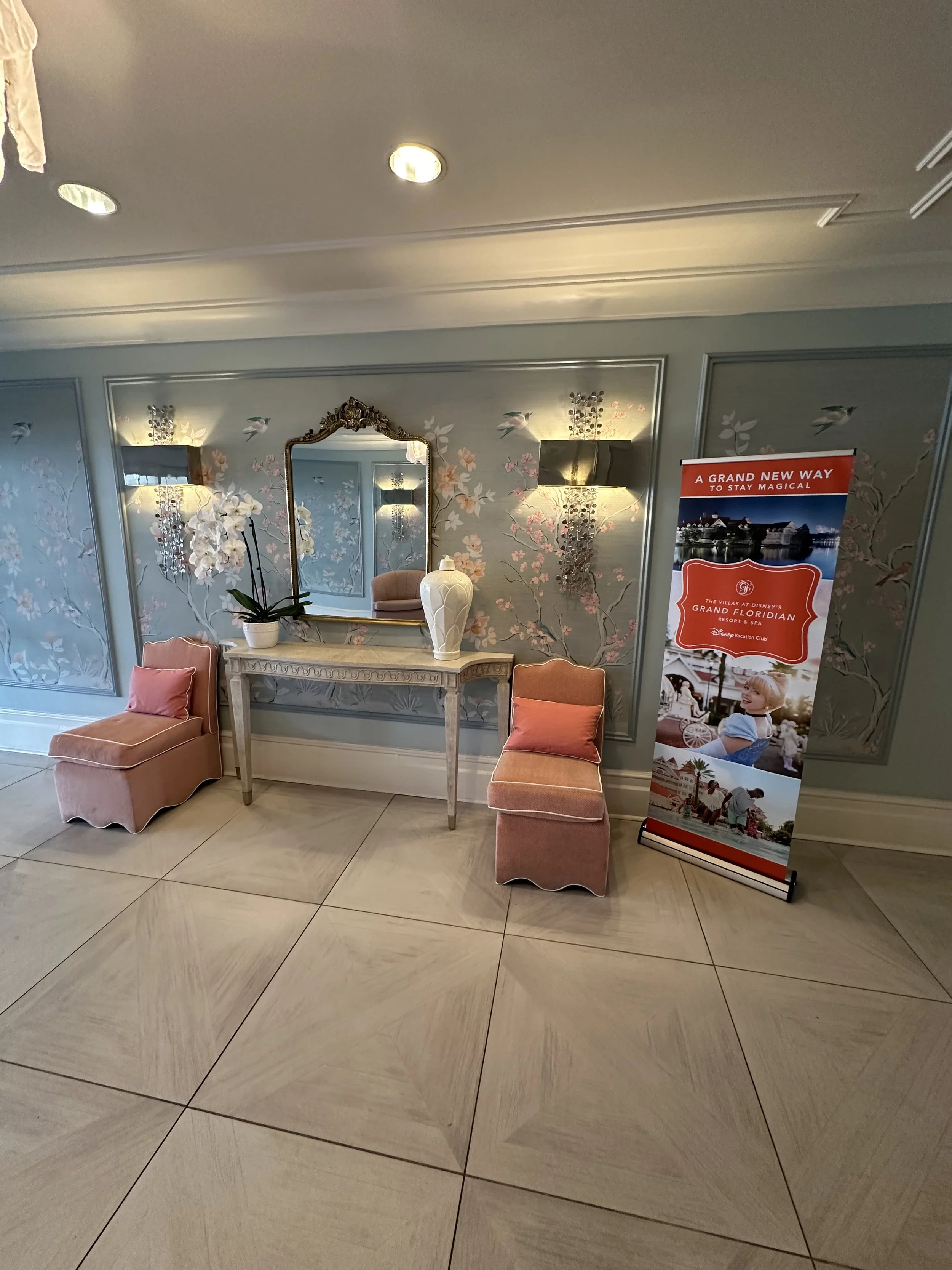 Elegant hotel lobby with floral wallpaper, a mirror, a side table with a decorative vase and orchid, and two pink upholstered chairs with peach cushions. A promotional standee for Disney's Grand Floridian Resort & Spa is beside the chairs.