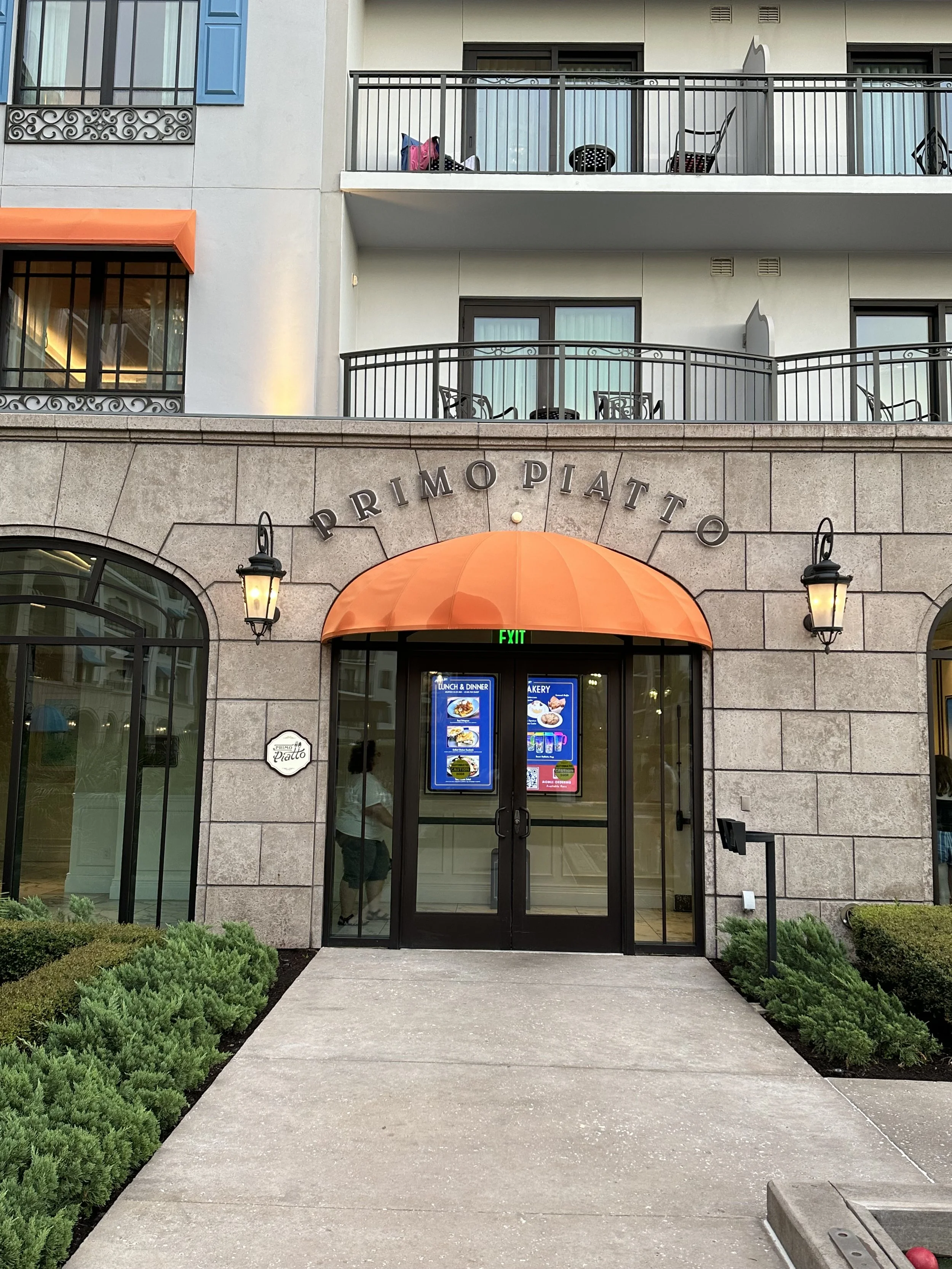 The entrance of a restaurant named 'Primo Piatt' with a stone facade, an orange awning, and black iron lanterns on either side of the door. There are signs for lunch, dinner, and bakery on the glass door, and greenery on both sides of a concrete walk