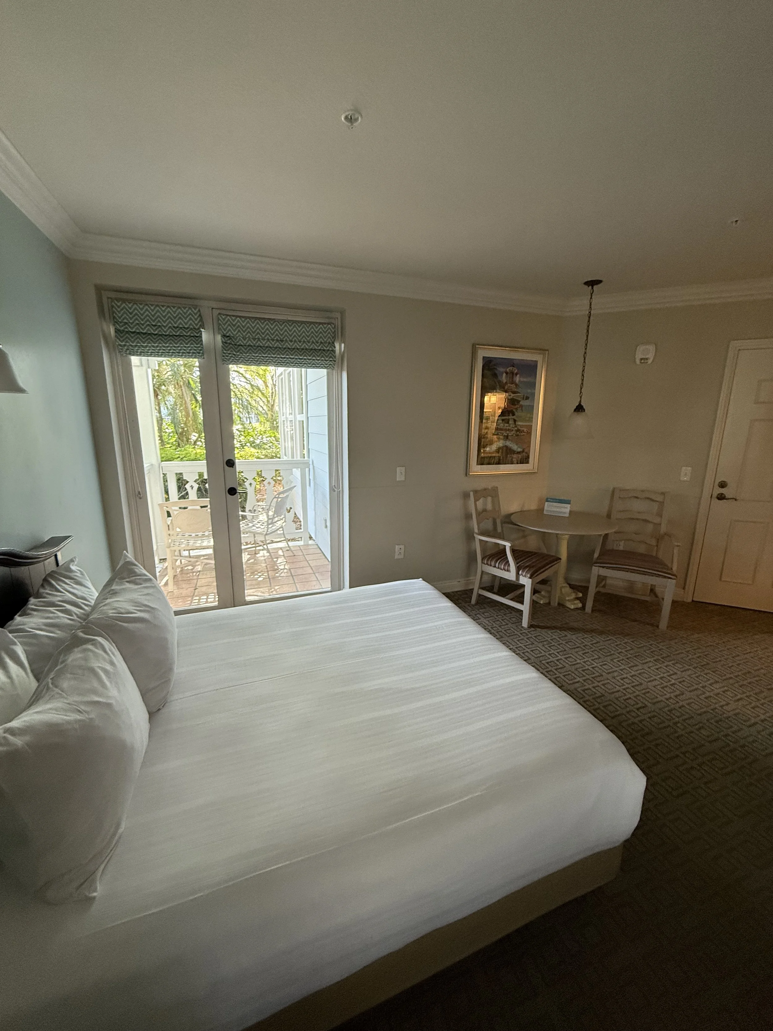 Hotel room with a neatly made bed, small balcony with outdoor seating, and a picture on the wall.
