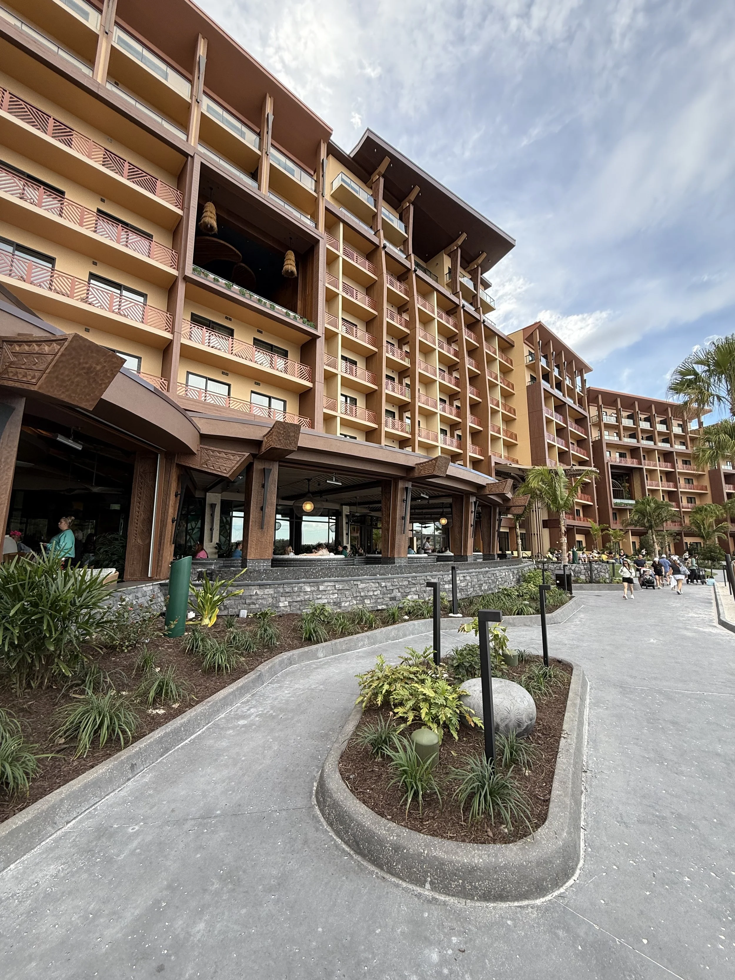 Exterior view of a multi-story hotel with balconies, a main entrance, and landscaping, including plants and palm trees, under a partly cloudy sky.