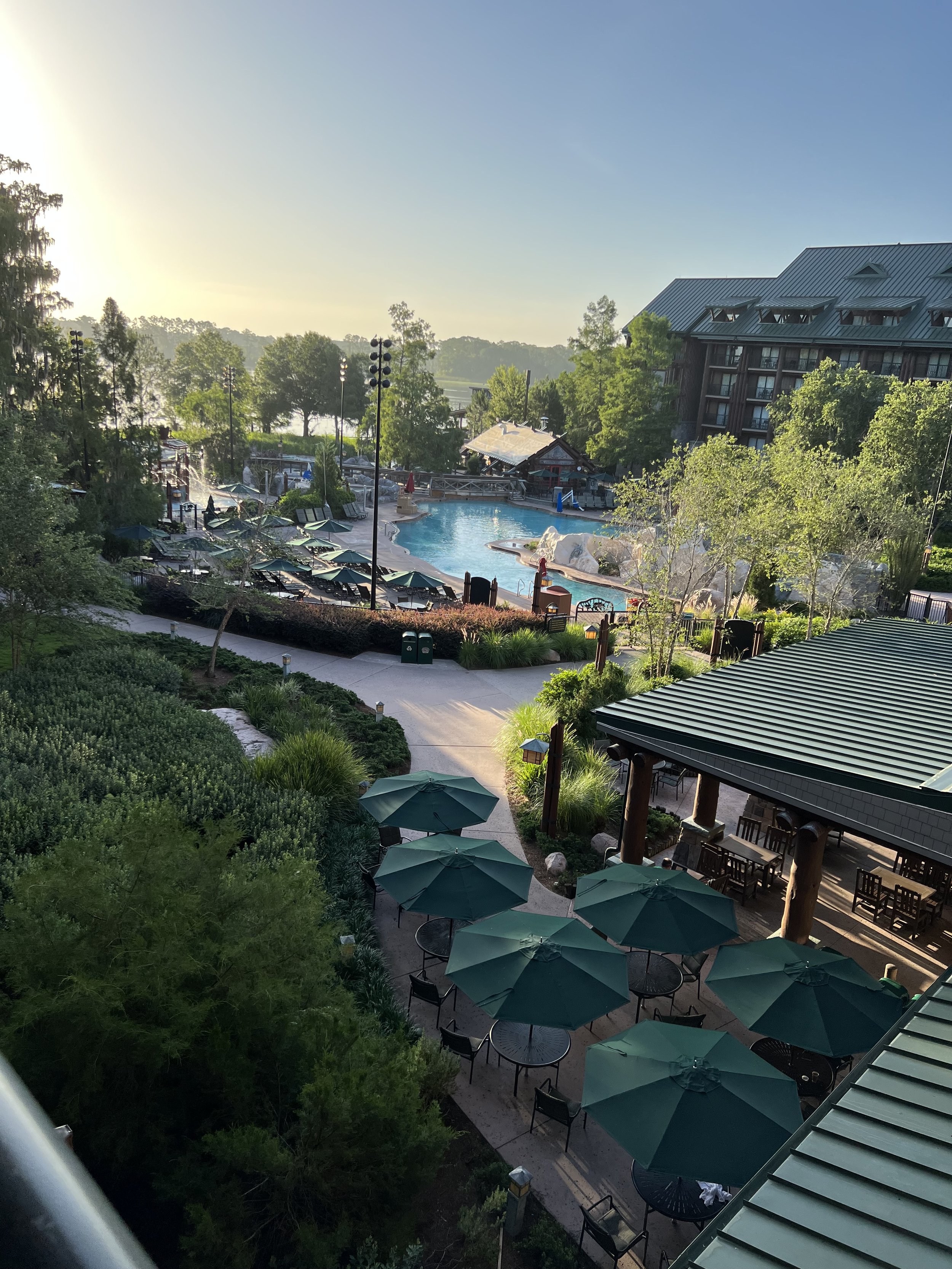 View of a resort pool area with shaded seating, umbrellas, lush greenery, and a modern hotel building in the background during sunrise or sunset.
