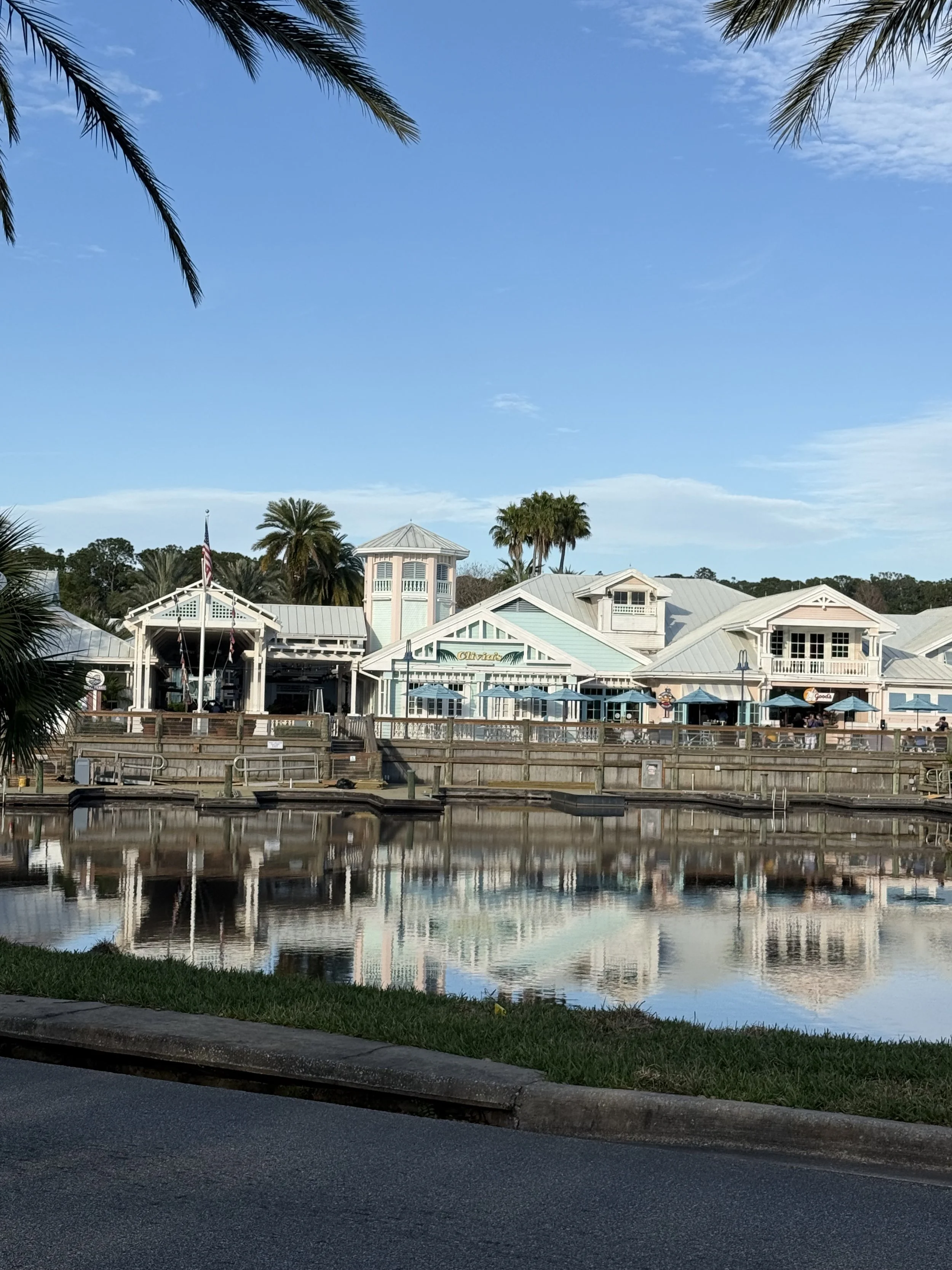 A pastel-colored waterfront building with outdoor seating and umbrellas, surrounded by palm trees under a blue sky, with its reflection in the water.