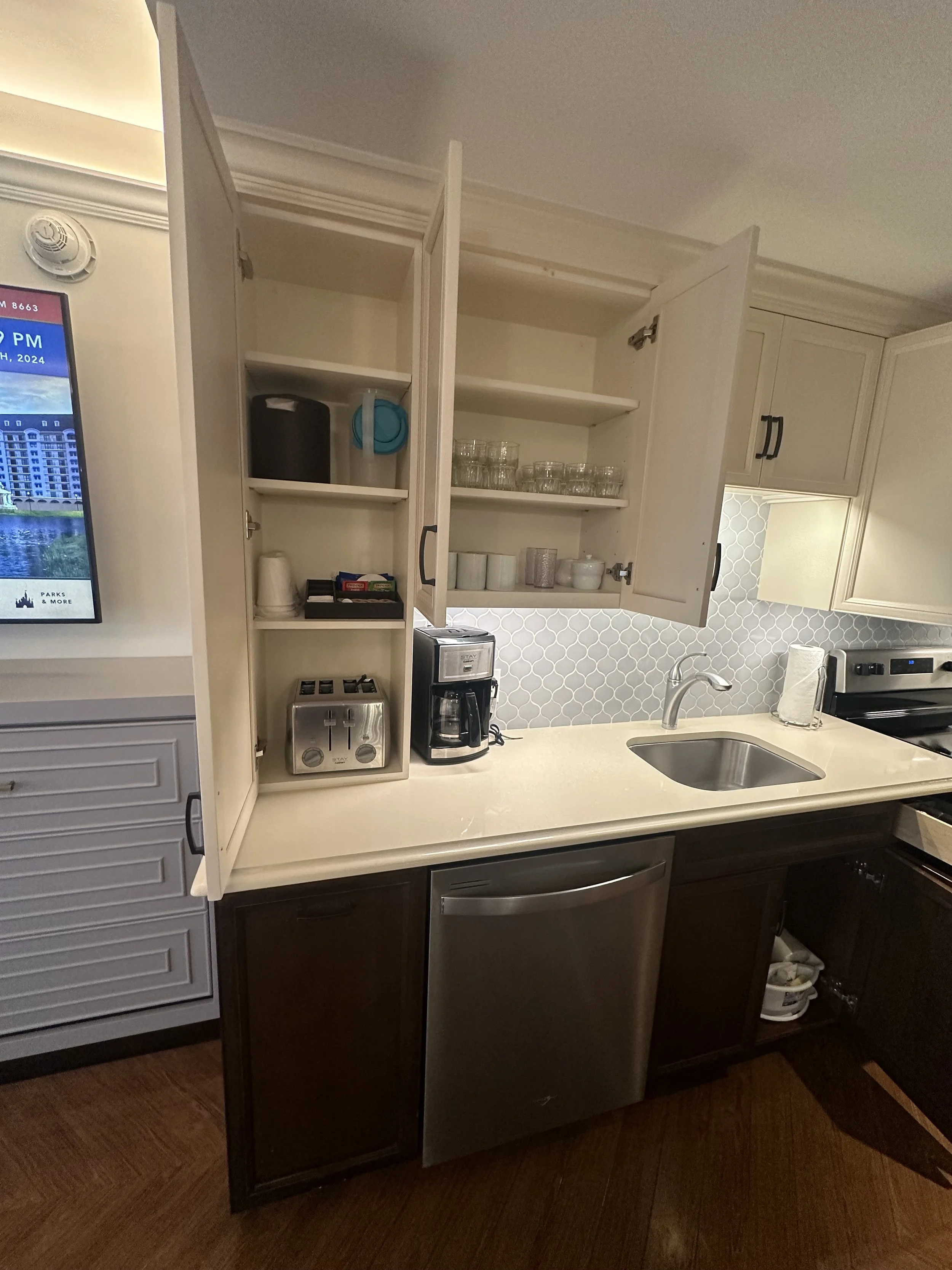 Open kitchen cabinet above the countertop with glasses, cups, and storage containers inside. Countertop has a coffee maker, toaster, and paper towel holder. Dishwasher below, with cabinet doors and floor visible.
