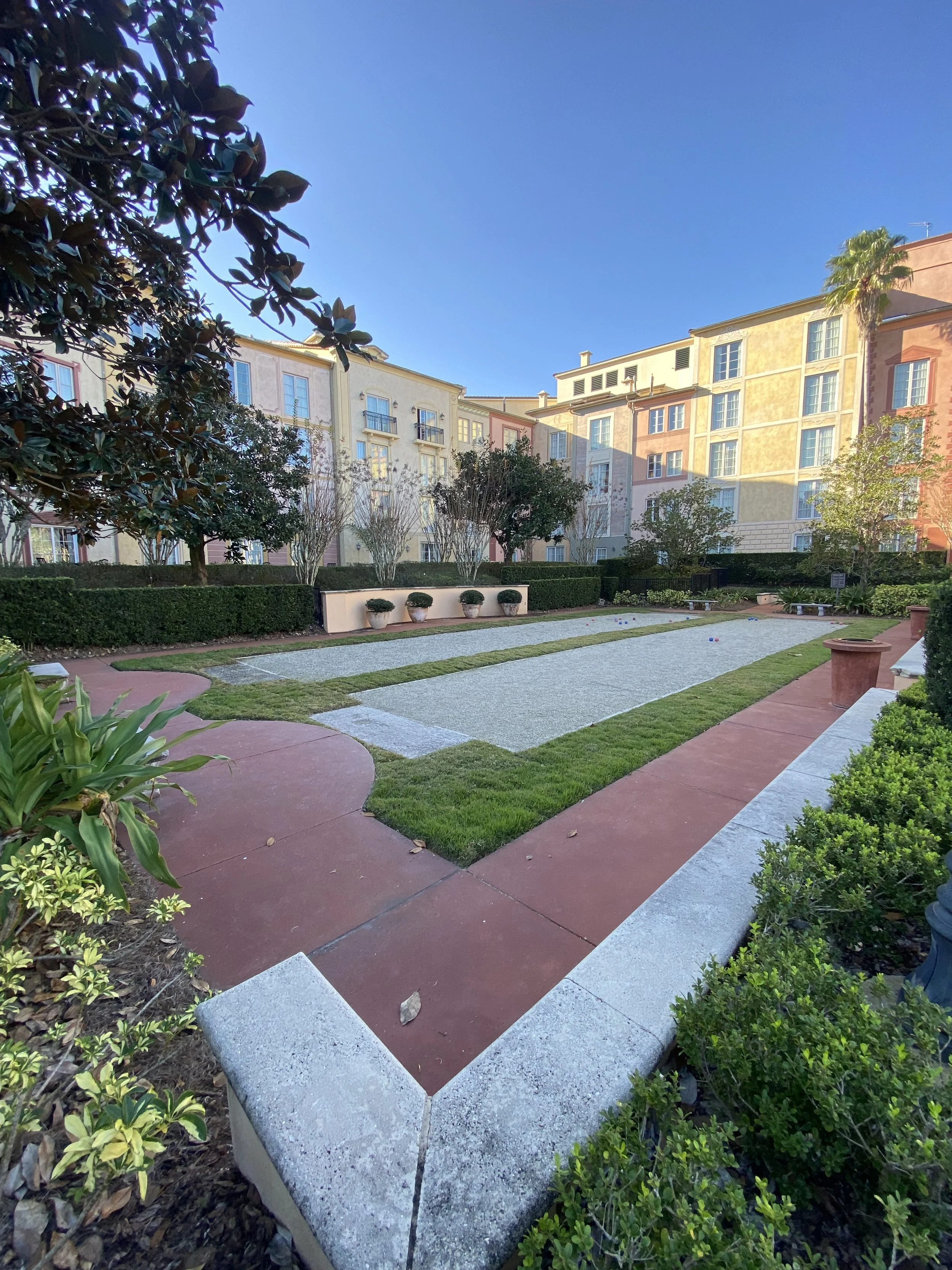 A courtyard with a lawn, gravel paths, and a bocce ball court, surrounded by apartment buildings and trees, with clear blue sky overhead.