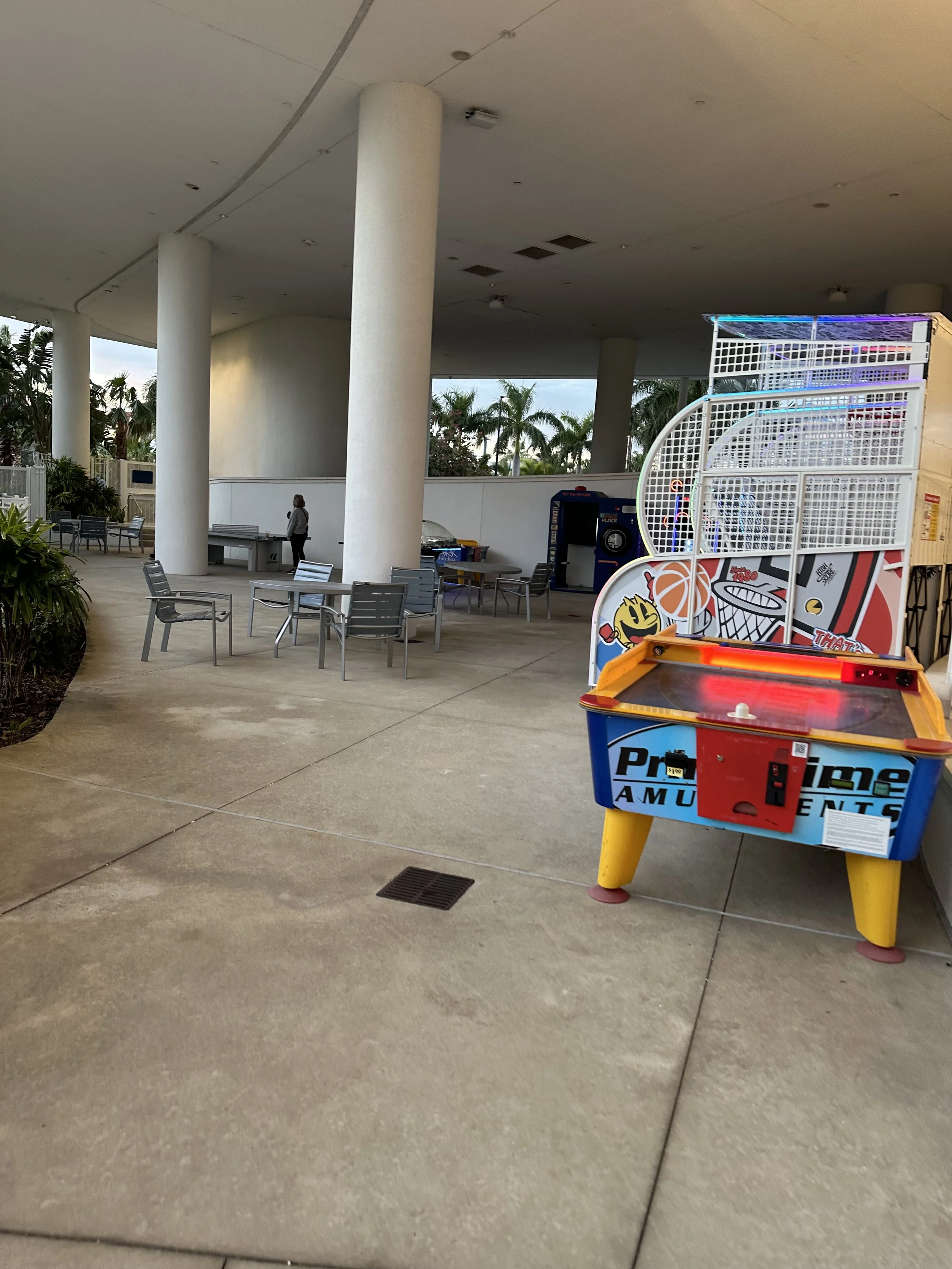 An outdoor area with tables and chairs under a roof, with a few people and arcade game machines, including a basketball game and a hockey game.