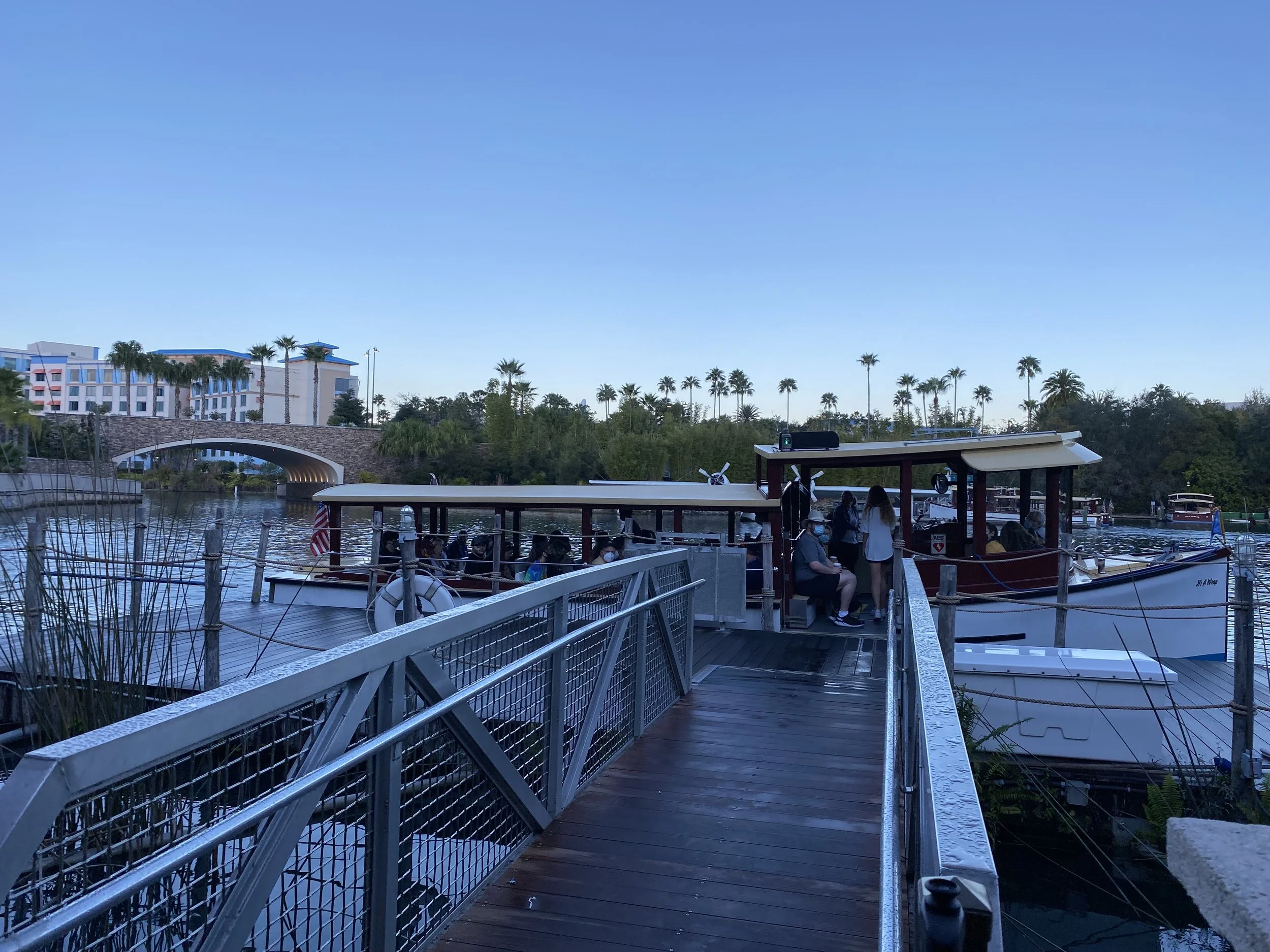People sitting on a docked boat at a marina during daytime with trees, a bridge, and buildings in the background.