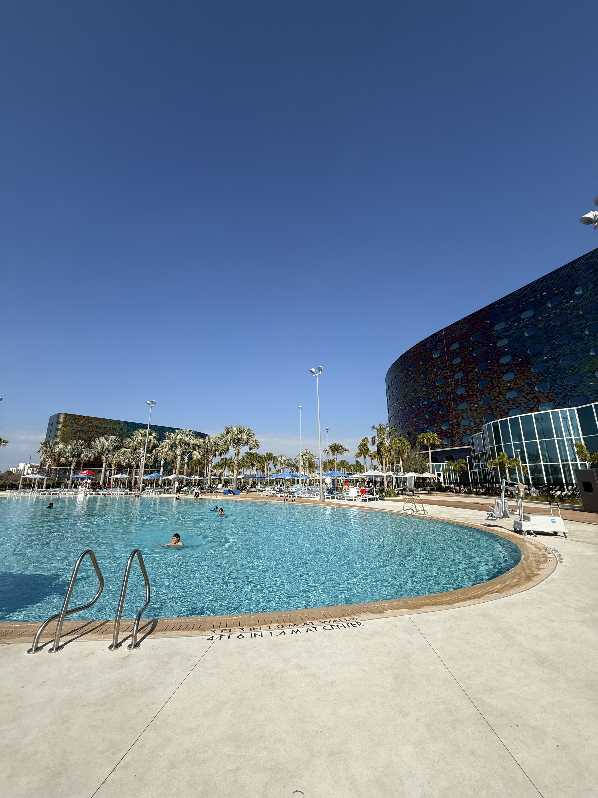 A large outdoor swimming pool with a few people swimming, surrounded by palm trees and lounge chairs under umbrellas. In the background, there are modern buildings with a dark exterior and many windows, set against a clear blue sky.