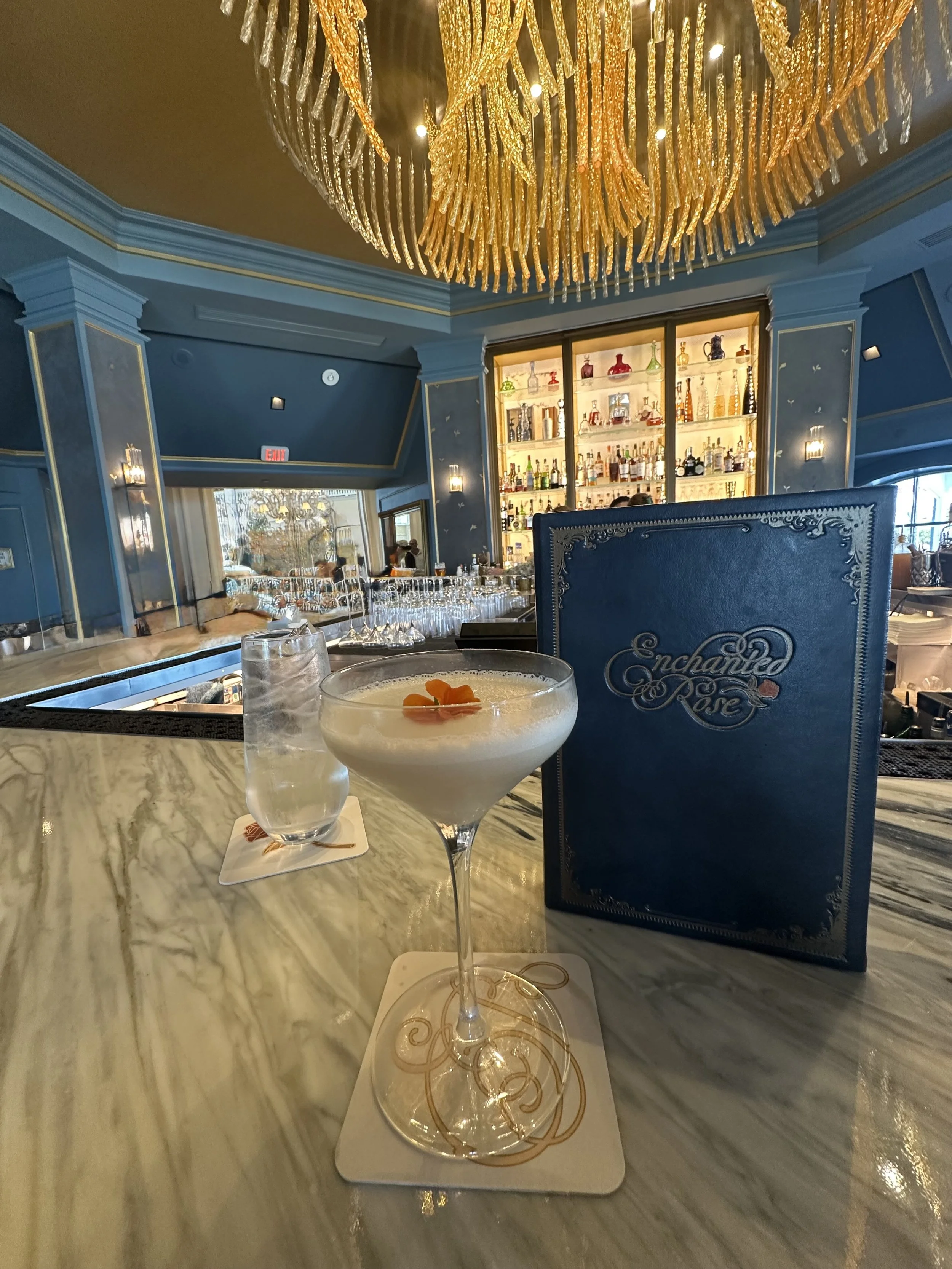 A cocktail glass with a frothy white drink garnished with orange peel on a marble bar counter, a blue Enchanted Rose menu, and a bar with bottles in the background, under a large gold chandelier.