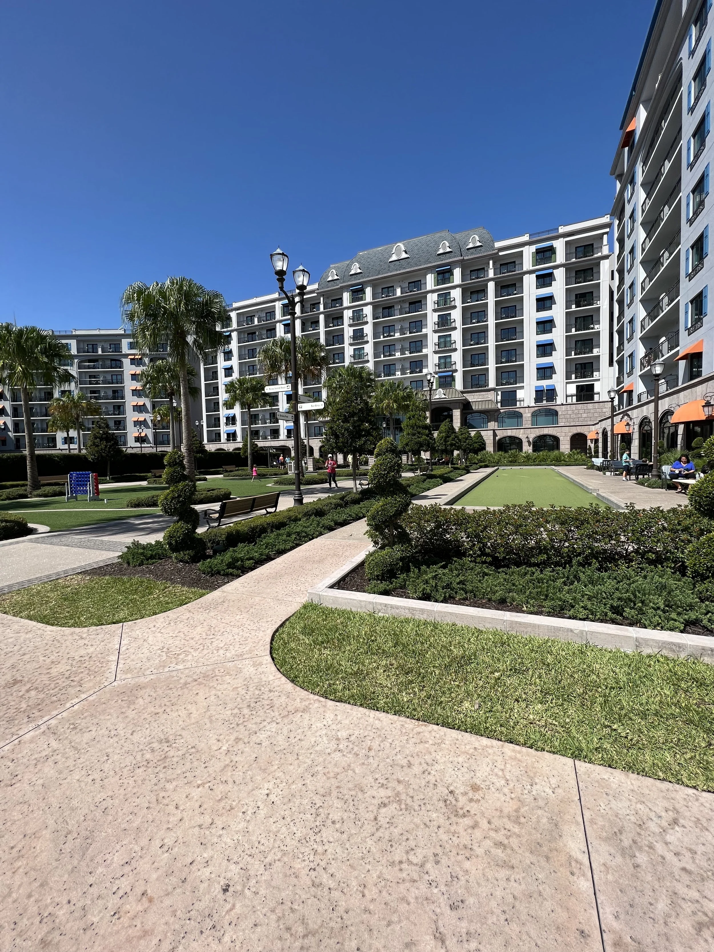 Outdoor view of a modern apartment complex with a landscaped courtyard featuring walkways, benches, trimmed bushes, and palm trees under a clear blue sky.