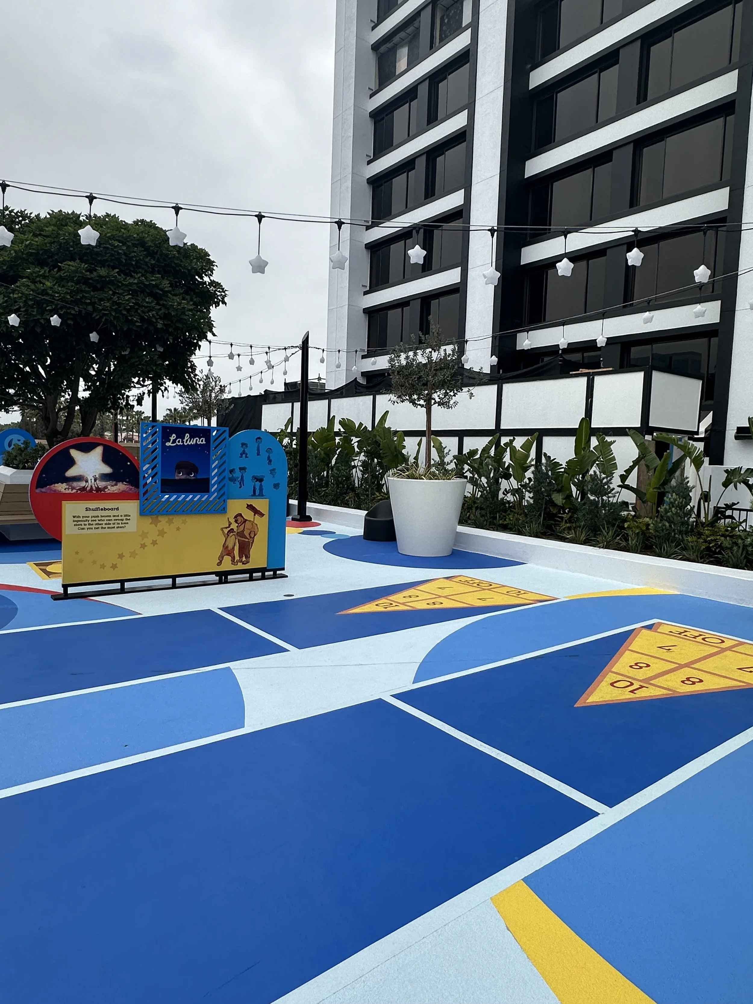 Outdoor recreational area with blue painted flooring resembling a hopscotch and shuffleboard game, decorated with planters, string lights, and a multi-story building in the background.