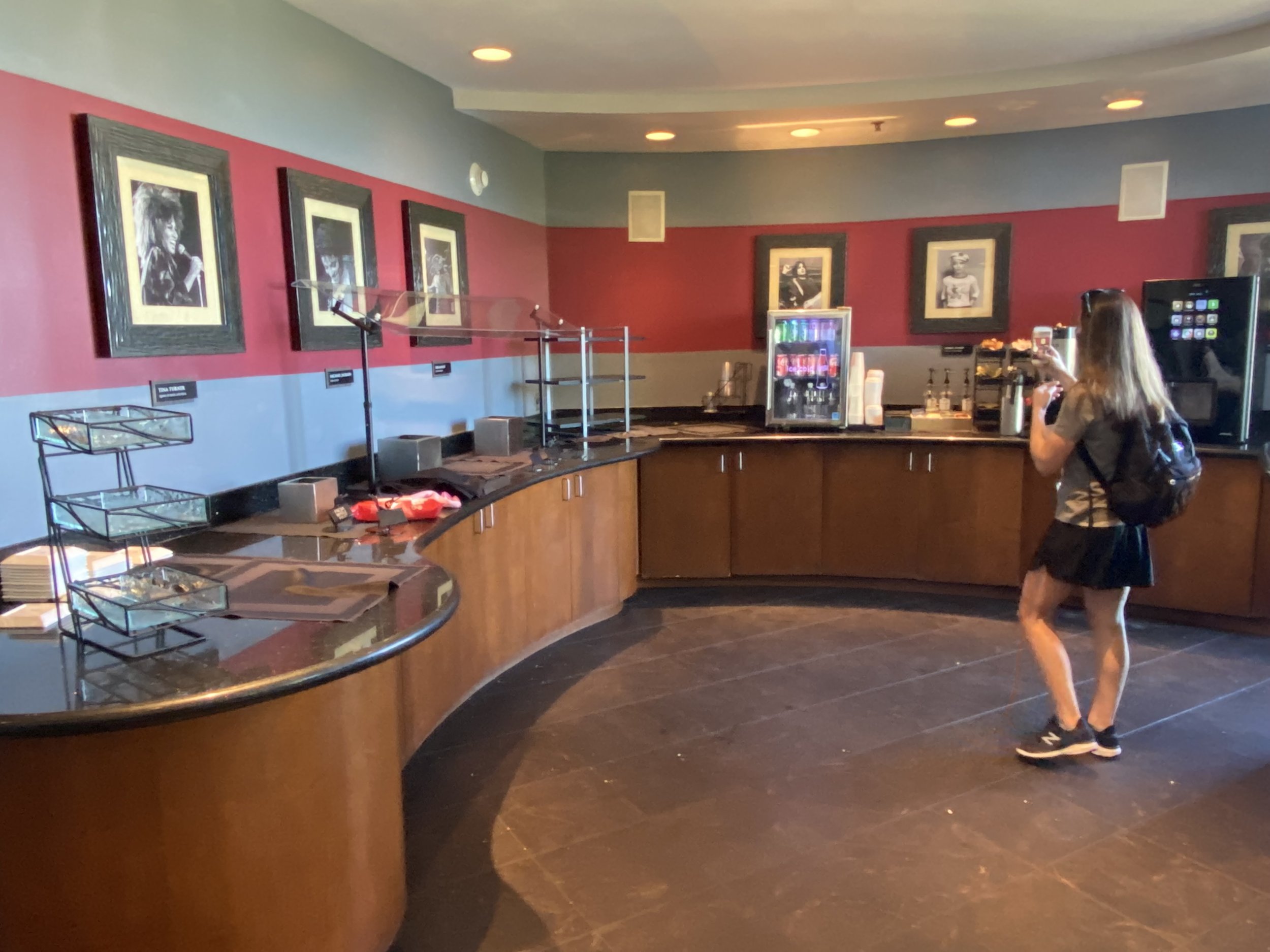 A woman in casual attire with a backpack is standing at a self-serve beverage station, taking a photo with her phone. The station has a mini fridge, dispenser, and framed black-and-white photographs on the wall behind it.