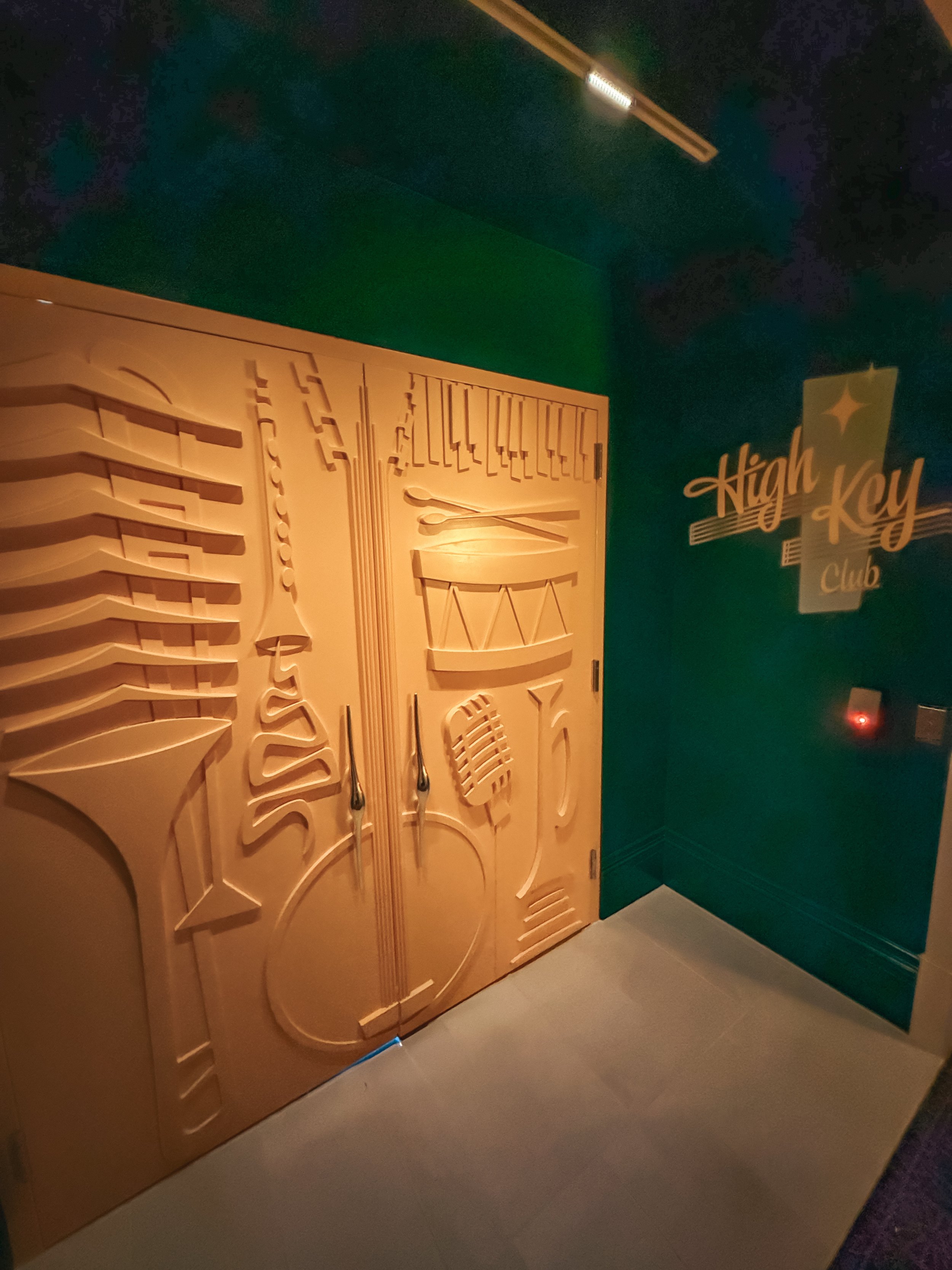 Decorative door with musical instruments and notes carved in relief, located in a room with High Key Club signage on dark green wall.
