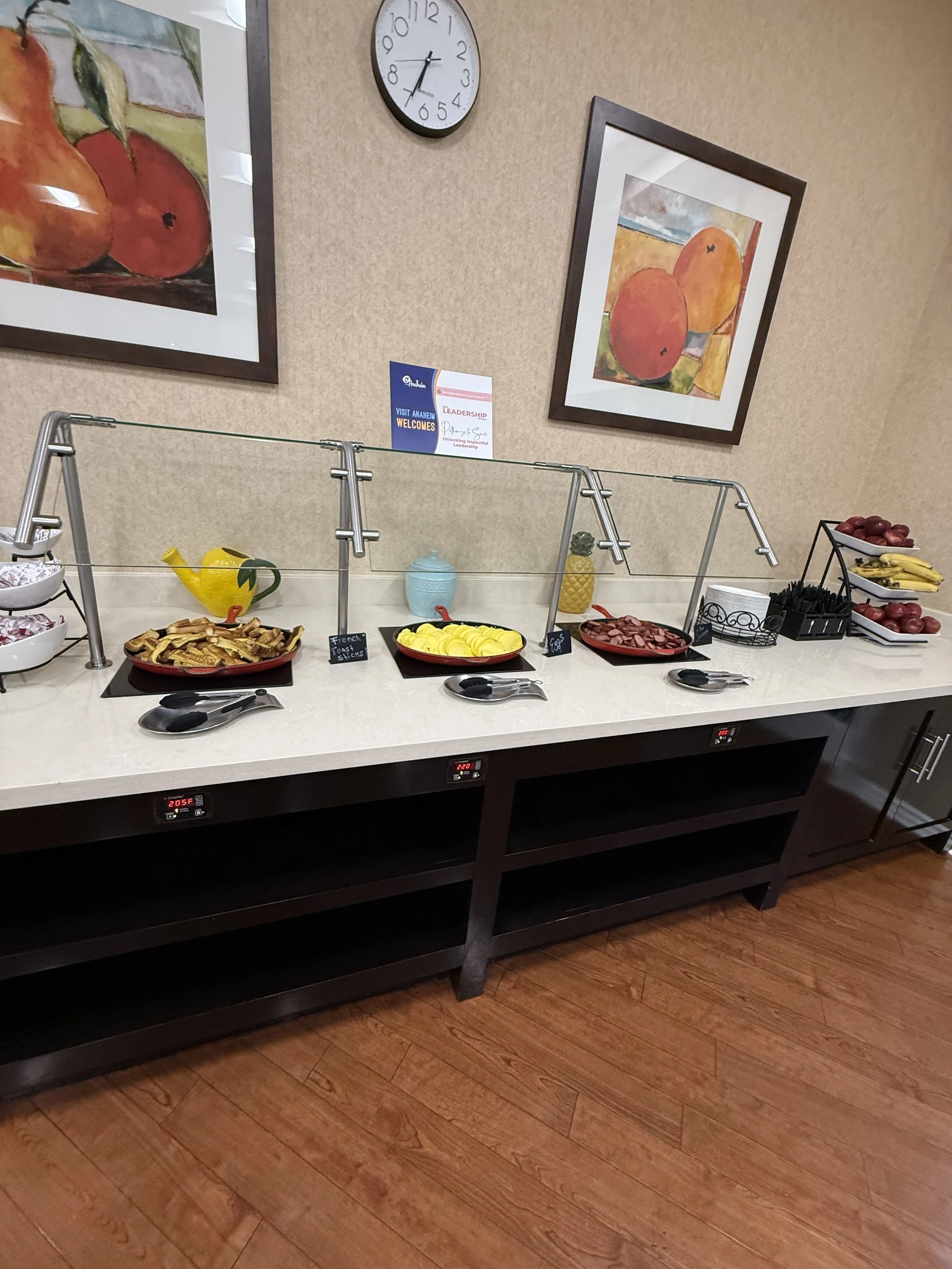 Buffet table with breakfast items including sliced turkey, sliced yellow peppers, grapes, bananas, and toast, with fridge temperature displays below and framed fruit artwork on the wall behind.