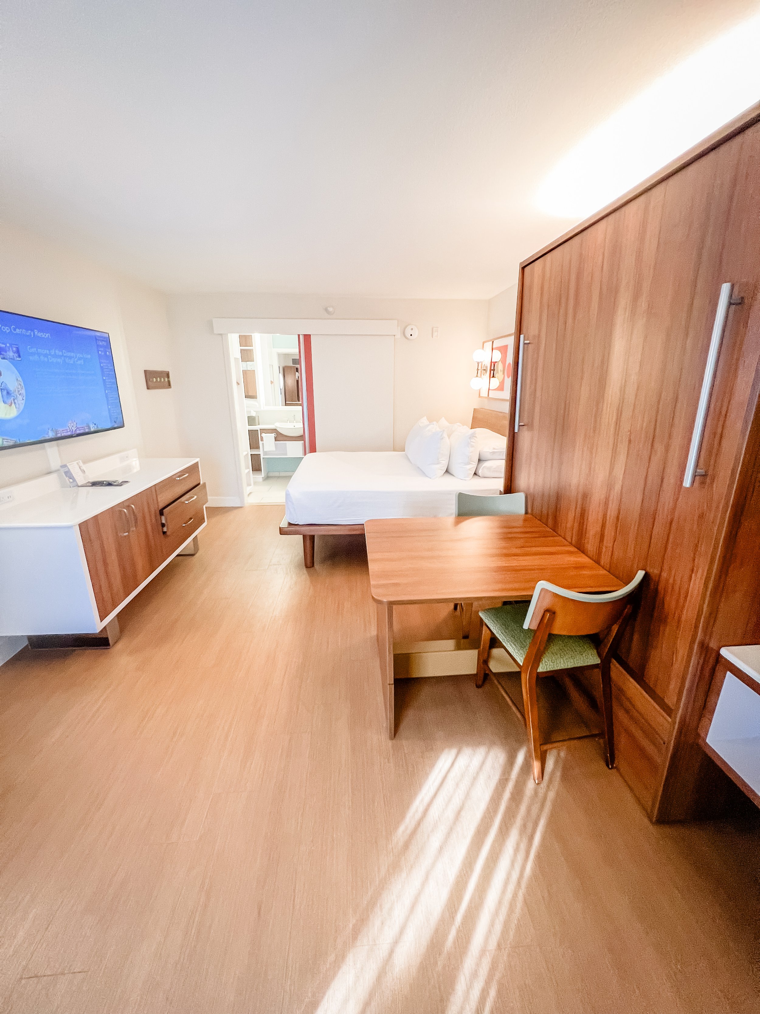 Hotel room with a bed, wooden furniture, a television, and a small desk with a chair. There is a bathroom visible through an open door.