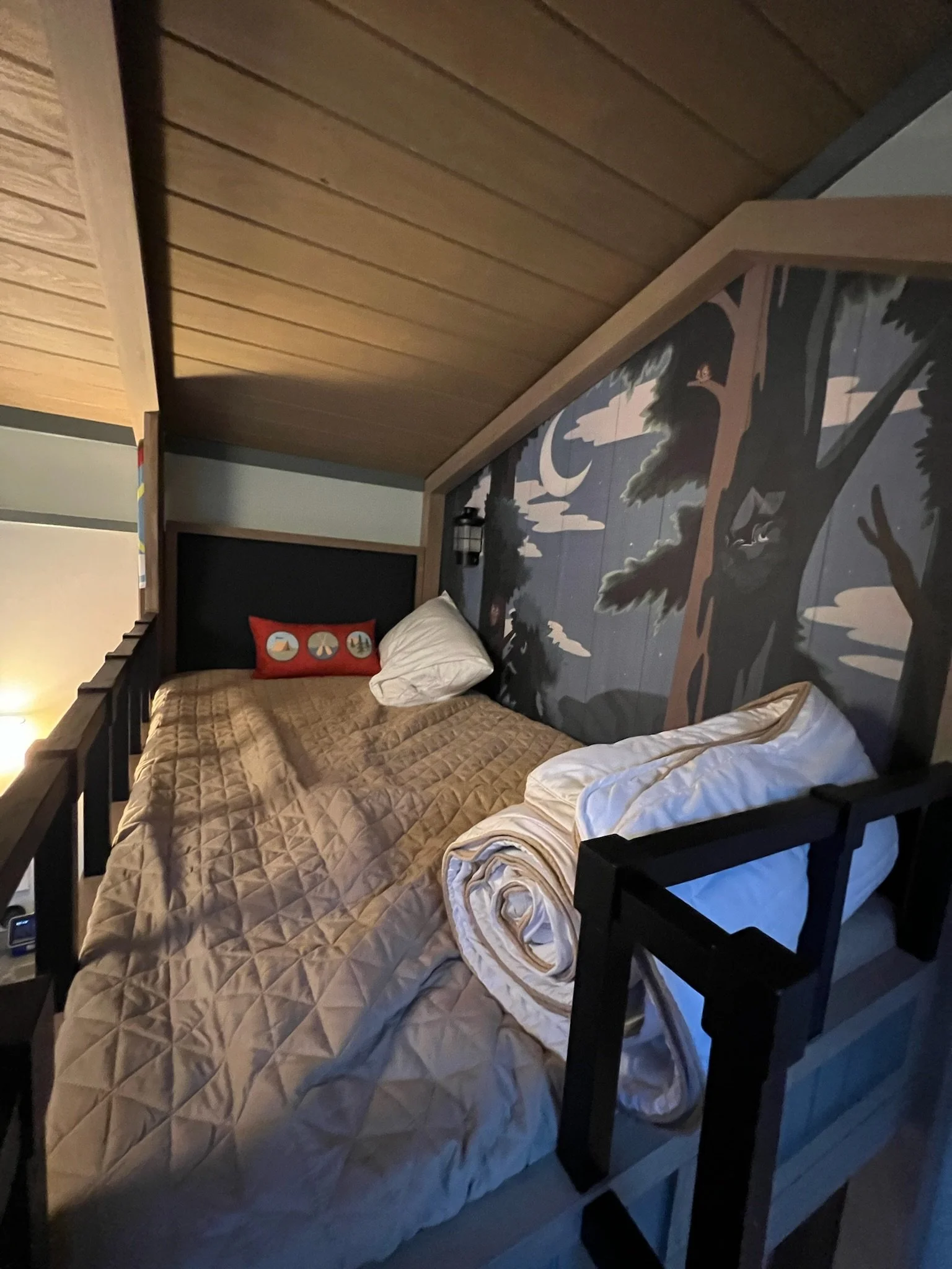 A cozy cabin bedroom with a wood-paneled ceiling, a mural of a forest on the wall, a beige quilted bedspread, a rolled-up white blanket, two white pillows, and a decorative pillow with camping symbols.