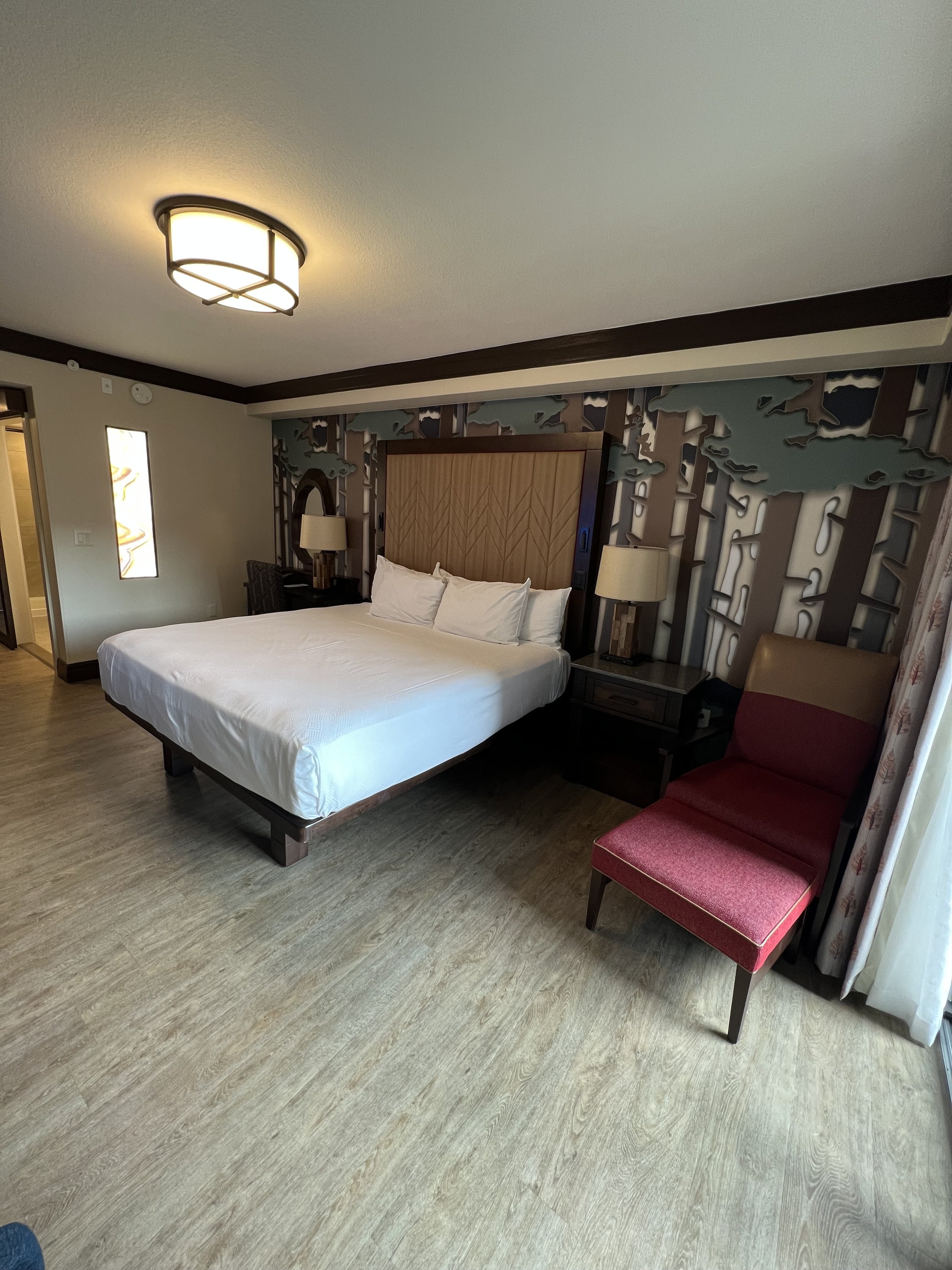 Hotel room with a large bed, decorative wall art behind the bed’s headboard, two bedside lamps, a red upholstered chair, and a window with curtains.