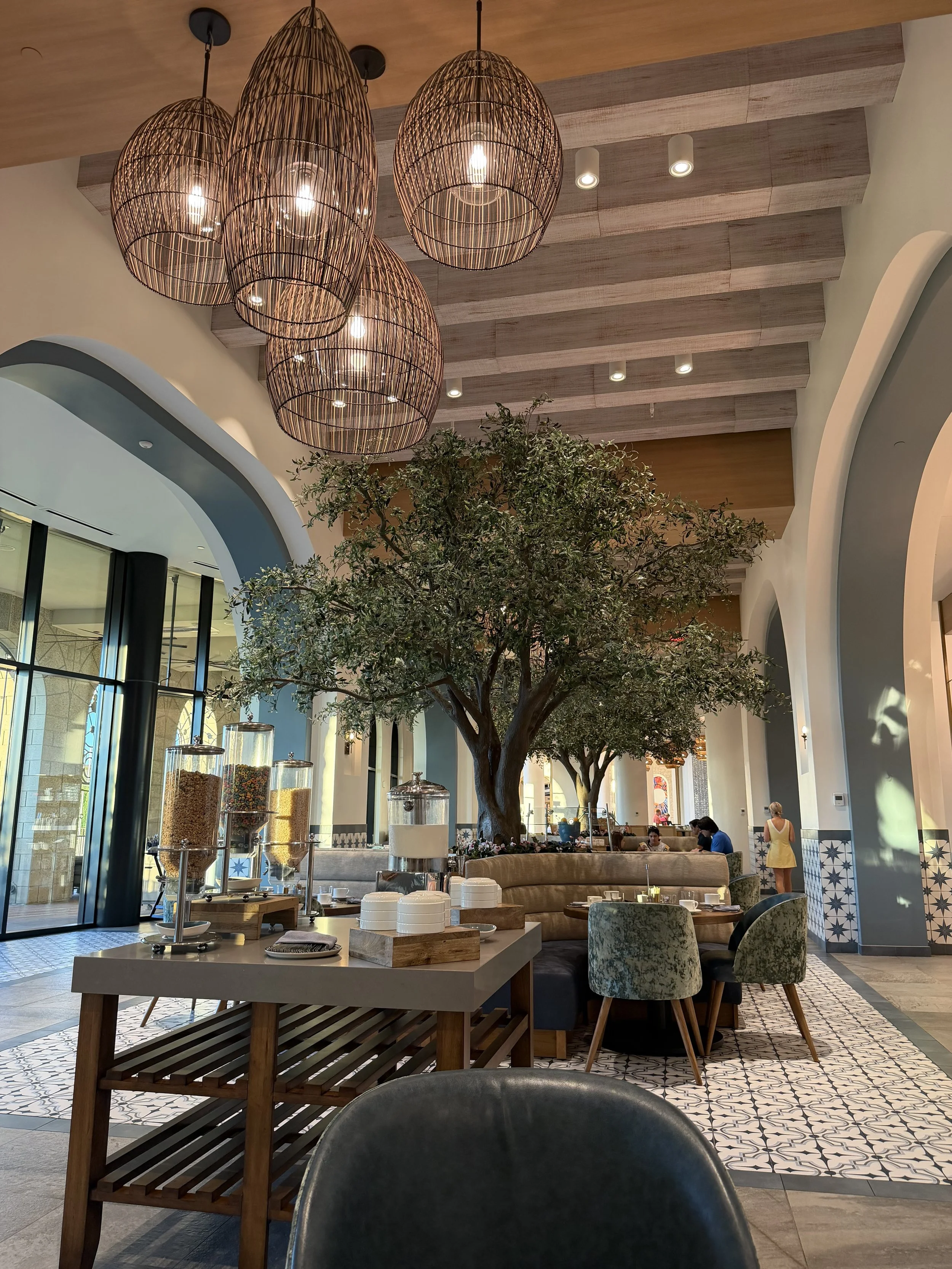 Elegant restaurant interior featuring large hanging wicker light fixtures, a large indoor tree, and a breakfast buffet with cereal dispensers.