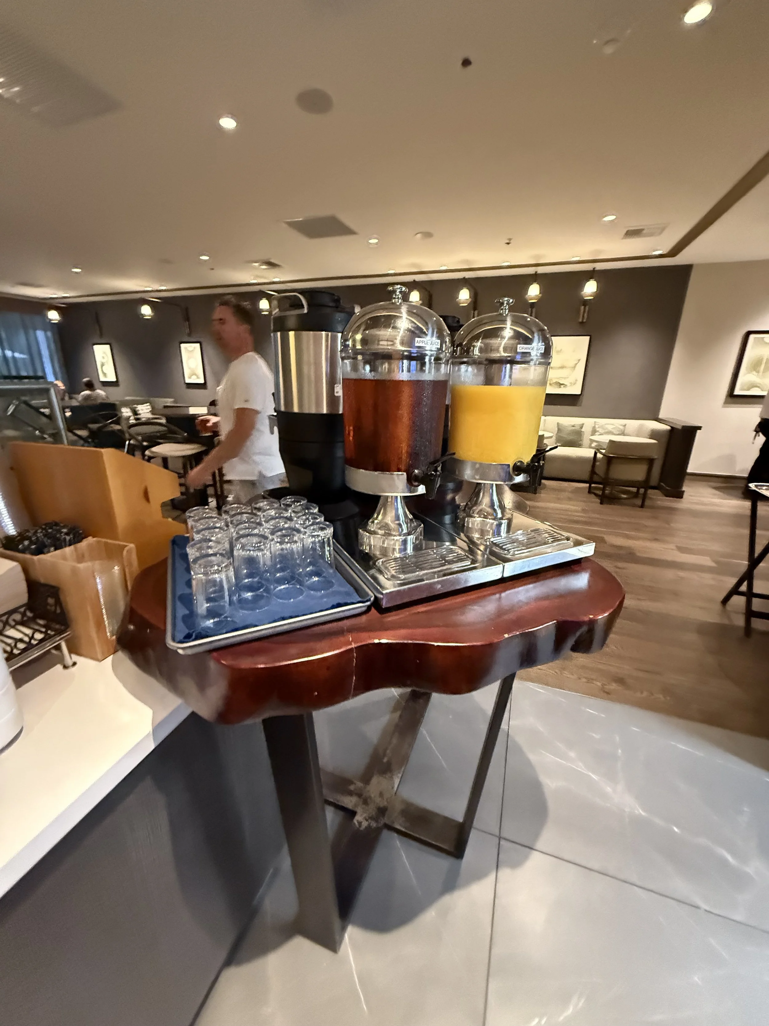 Coffee and juice station with dispensers of apple juice and orange juice, glasses, and a coffee urn in a restaurant setting.