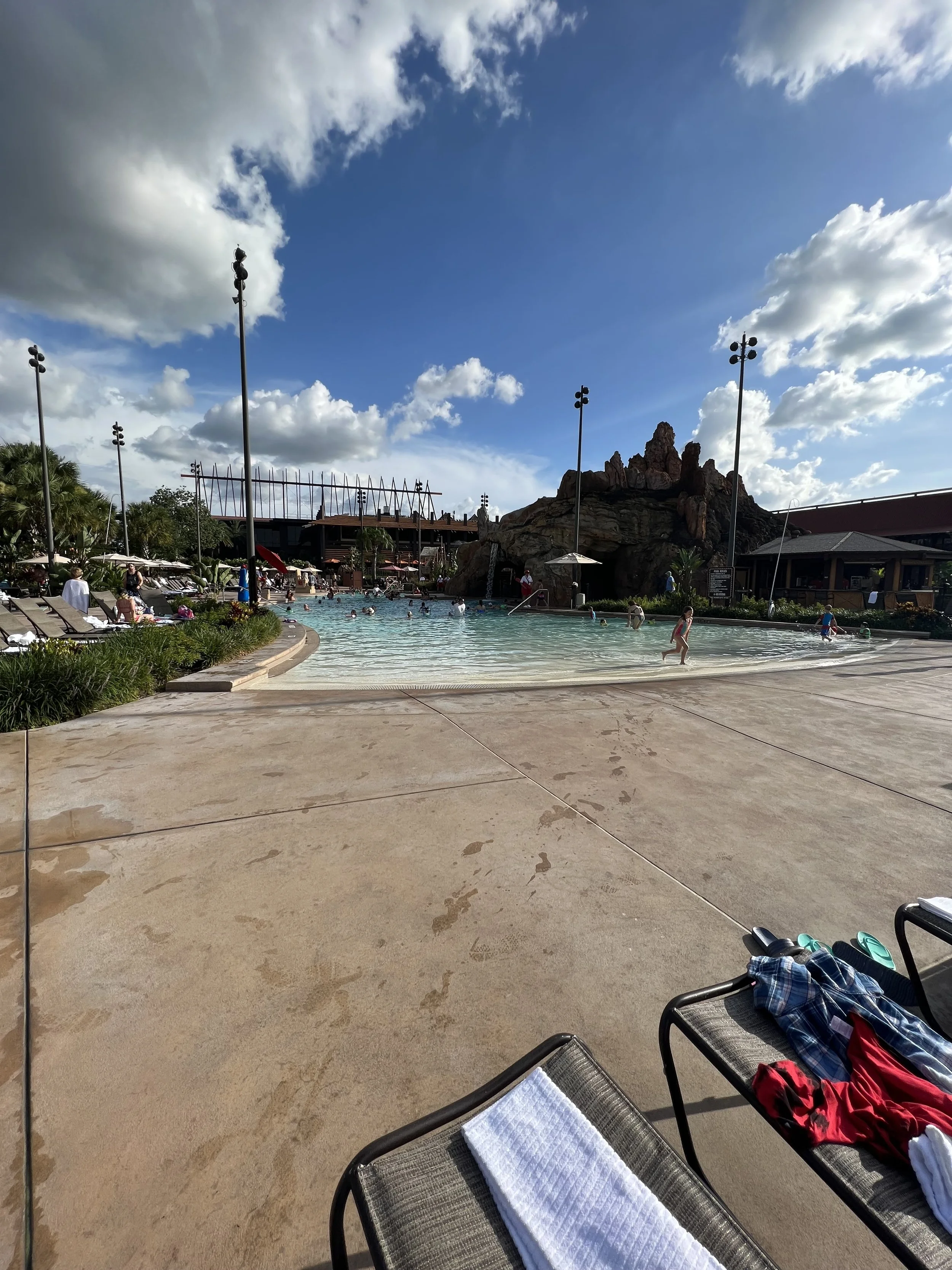 Poolside scene with lounge chairs and towels in the foreground, people swimming and playing in the pool, large rock formation, tropical plants, buildings, and cloudy sky overhead.