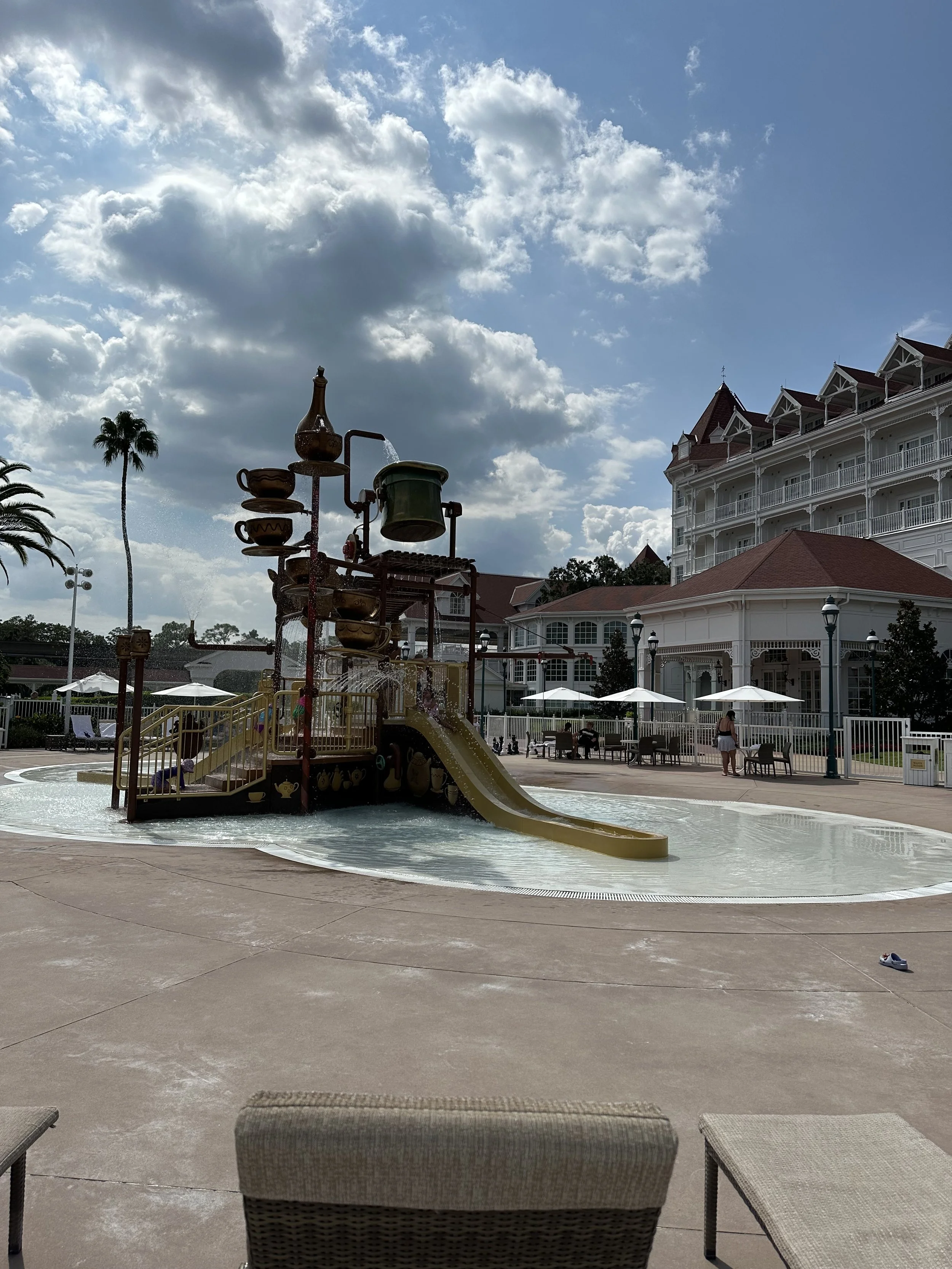 Empty water play area with a large boat-shaped spray structure, slide, and shallow water. In the background, there are white building with red roofs, umbrellas, trees, and a few people.