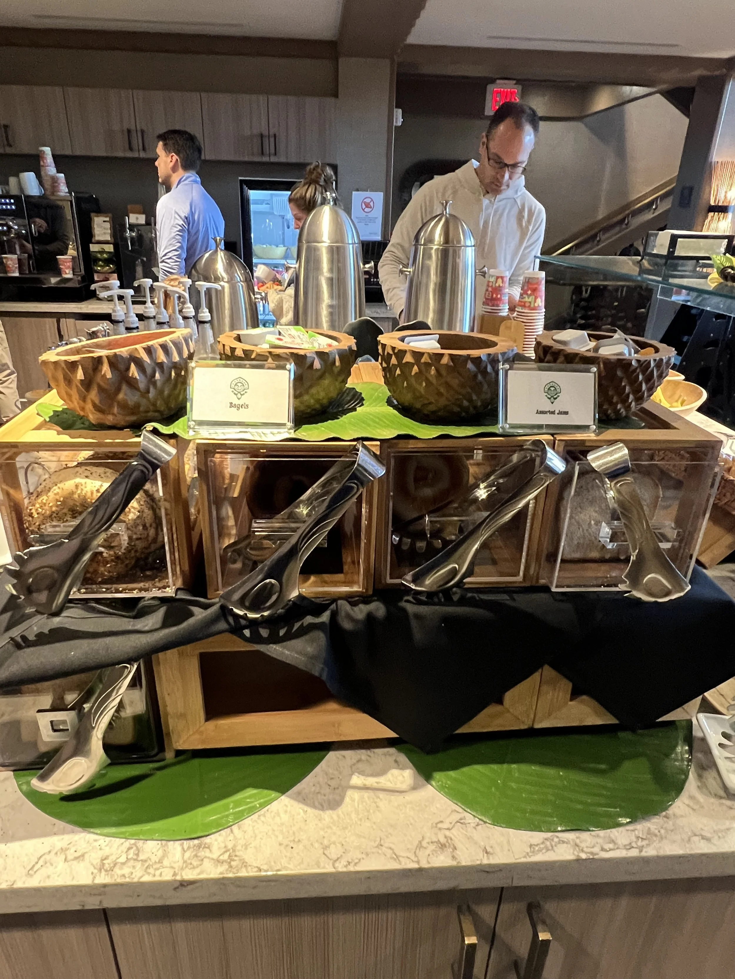 A breakfast buffet setup including bowls of bagels and assorted jams, with tongs for serving, in a restaurant or hotel lounge area.