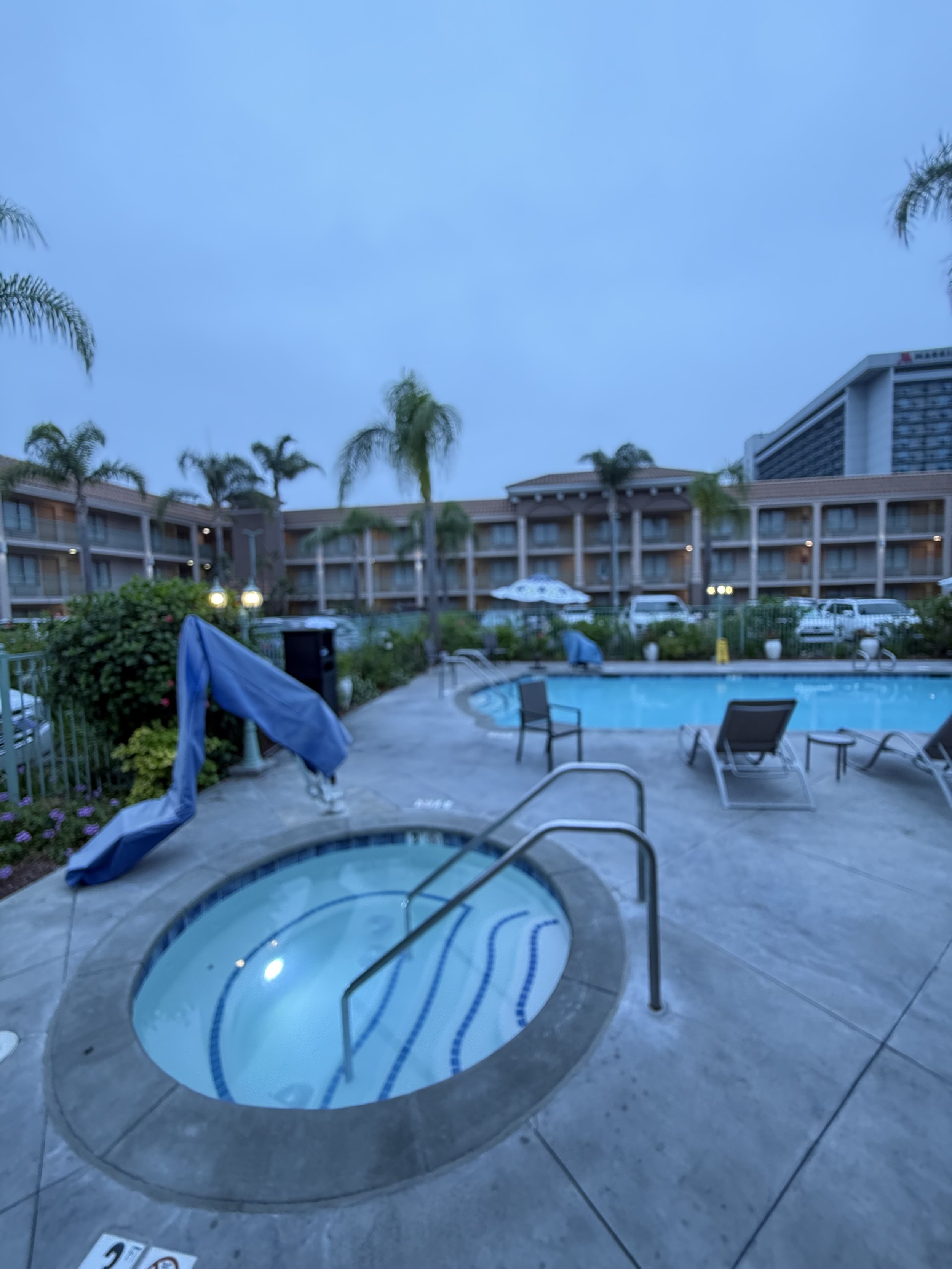 Empty swimming pool and hot tub area at a hotel during dusk, with lounge chairs and palm trees surrounding the pool.