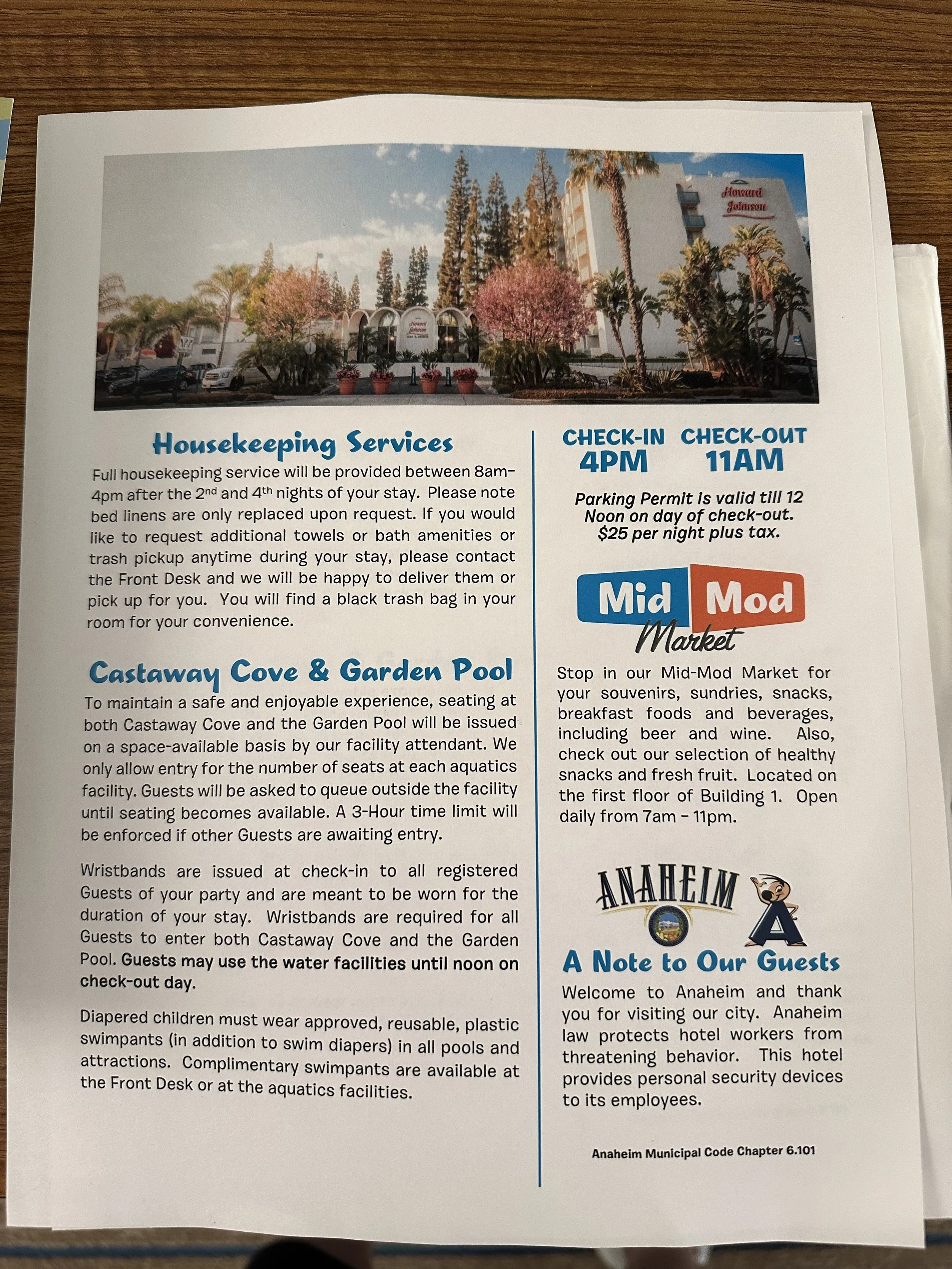 A printed informational sheet about hotel services, featuring a photo of a hotel with trees and a blue sky at the top. The text describes housekeeping, pool rules, a market, and guest welcome information for Anaheim.