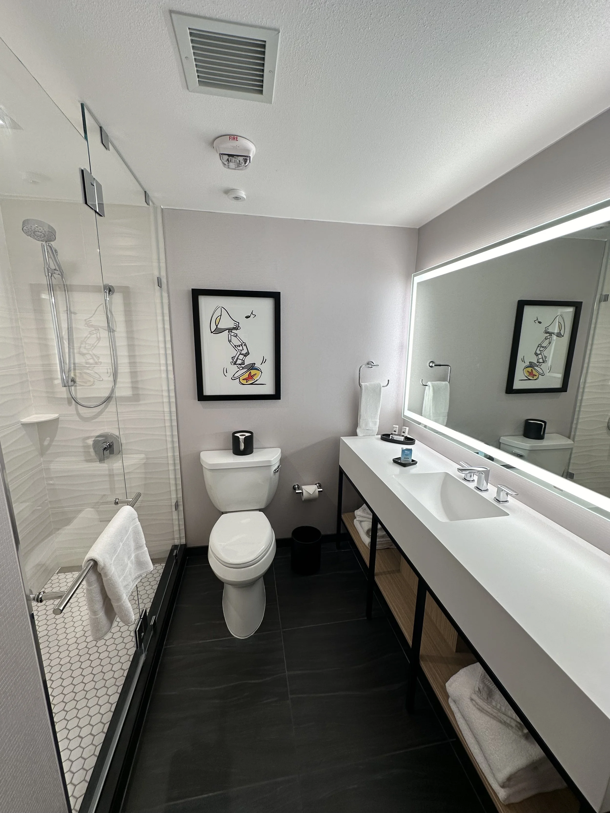 Modern hotel bathroom with a glass-enclosed shower, a white toilet, a black trash can, a large illuminated mirror above a white sink with a black frame. The vanity has open shelves with towels and toiletries, and there is a framed cartoon art on the 